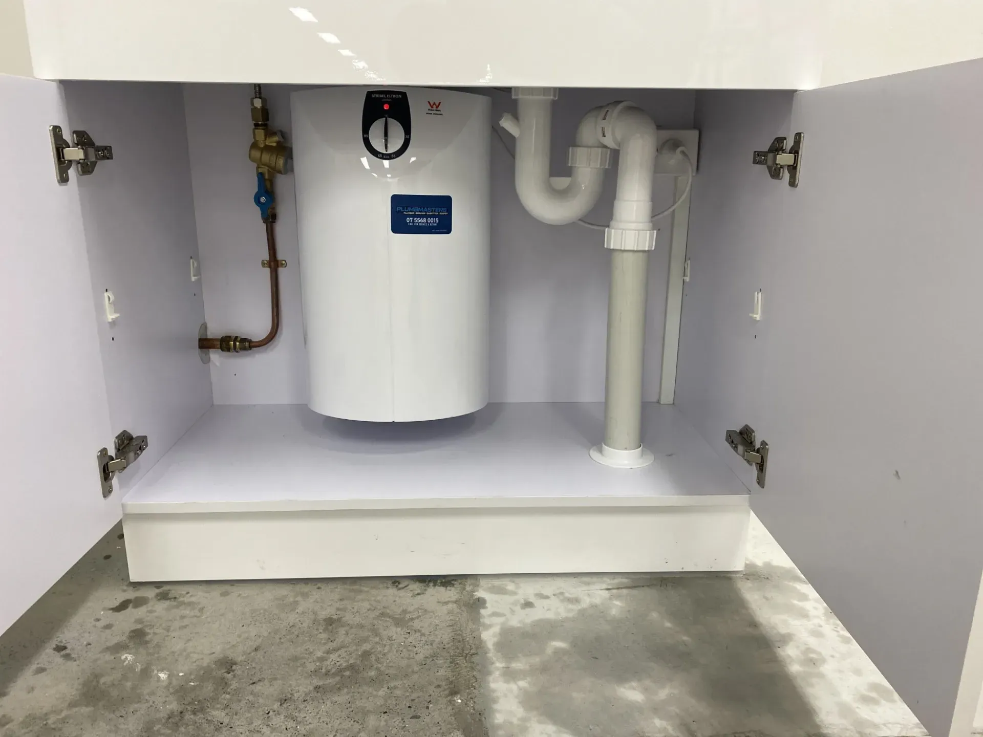 Under-sink cabinet with a white water heater and drain piping visible against a white interior. — Plumbmasters In Burleigh Heads, QLD