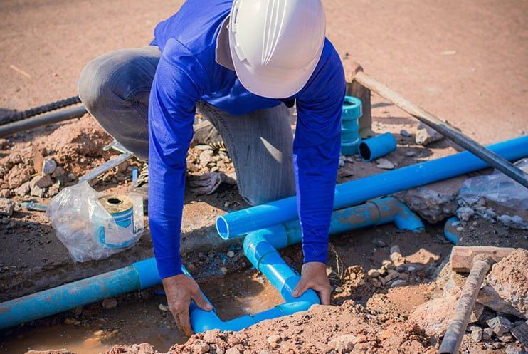 A Man Wearing A Hard Hat Is Working On A Pipe In The Dirt — Plumbmasters In Burleigh Heads, QLD