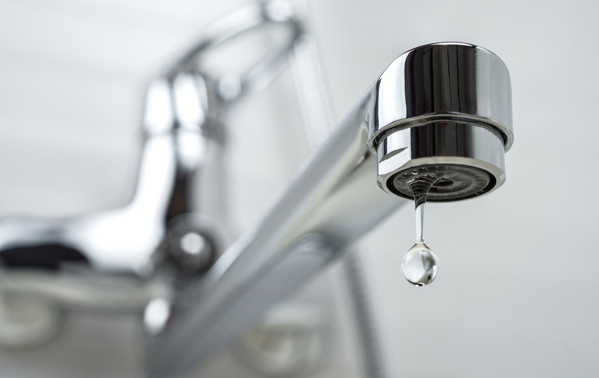 A chrome kitchen faucet dripping a single clear water droplet against a soft, out-of-focus background.