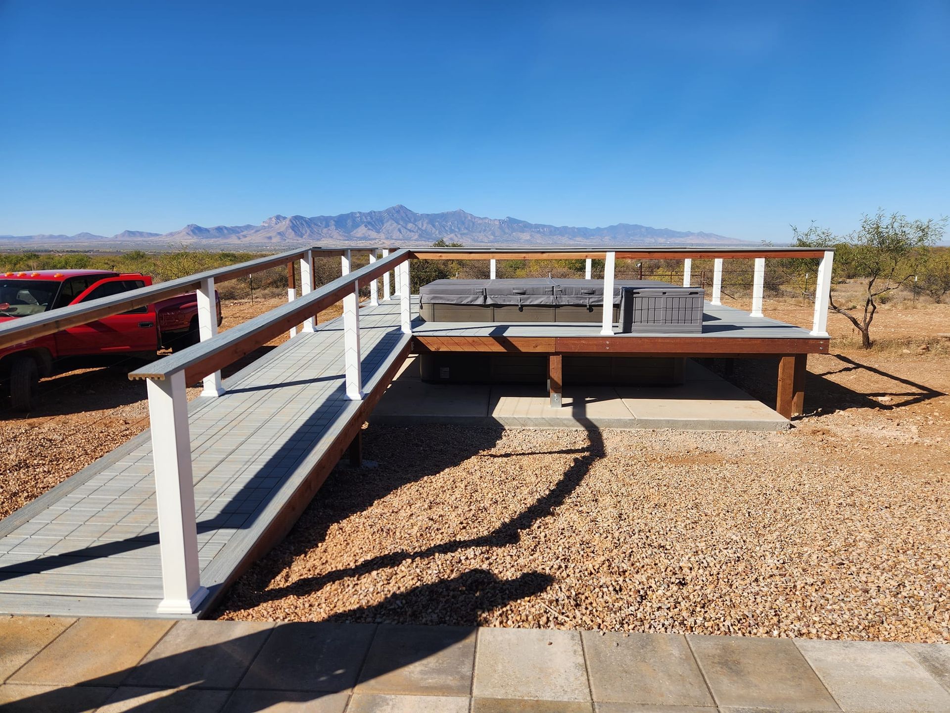 Wooden deck with ramp and hot tub, surrounded by desert landscape and mountains, under a blue sky.
