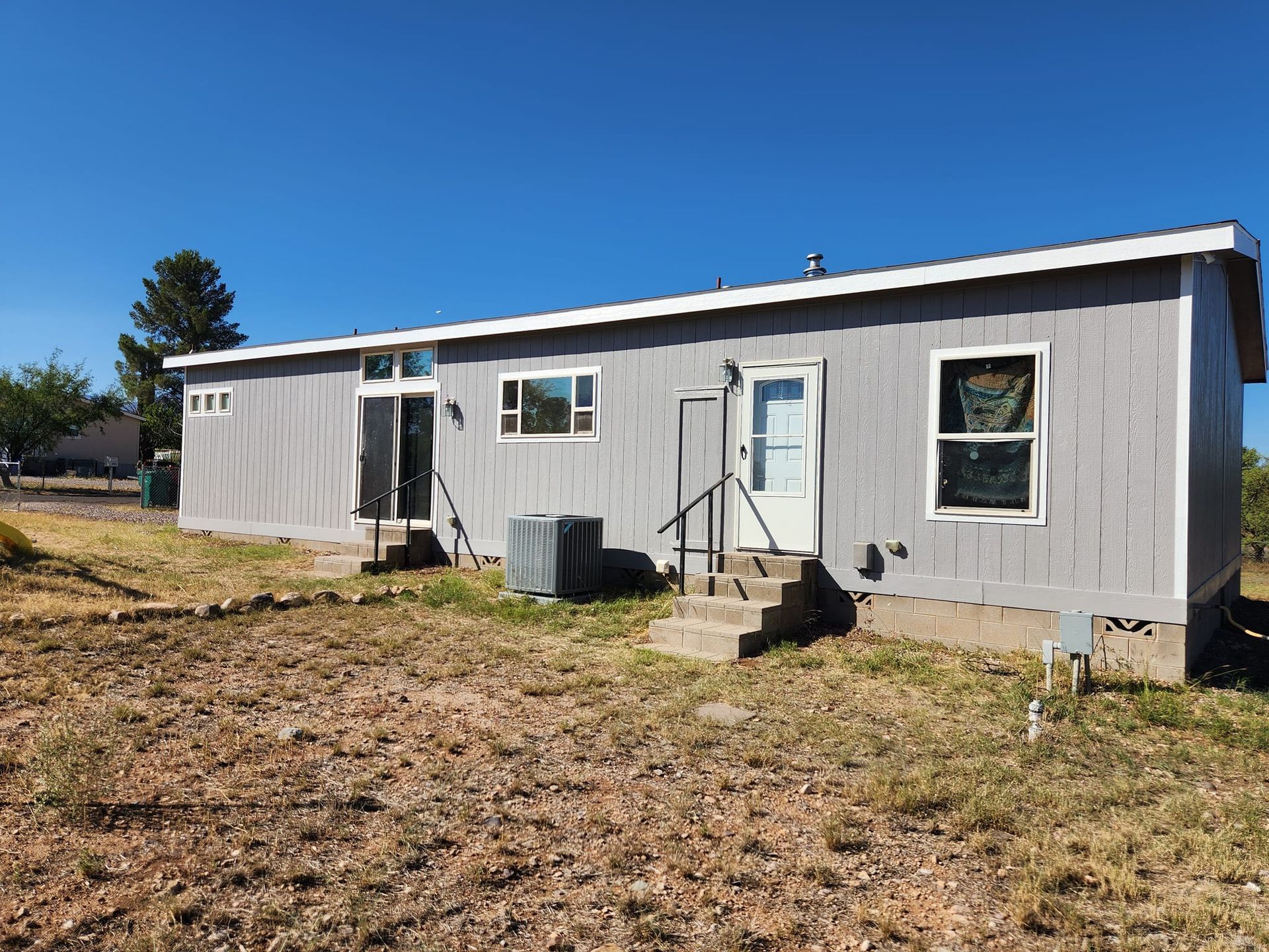 Back view of a gray, single-story house with a white door, windows, and steps; a clear blue sky.