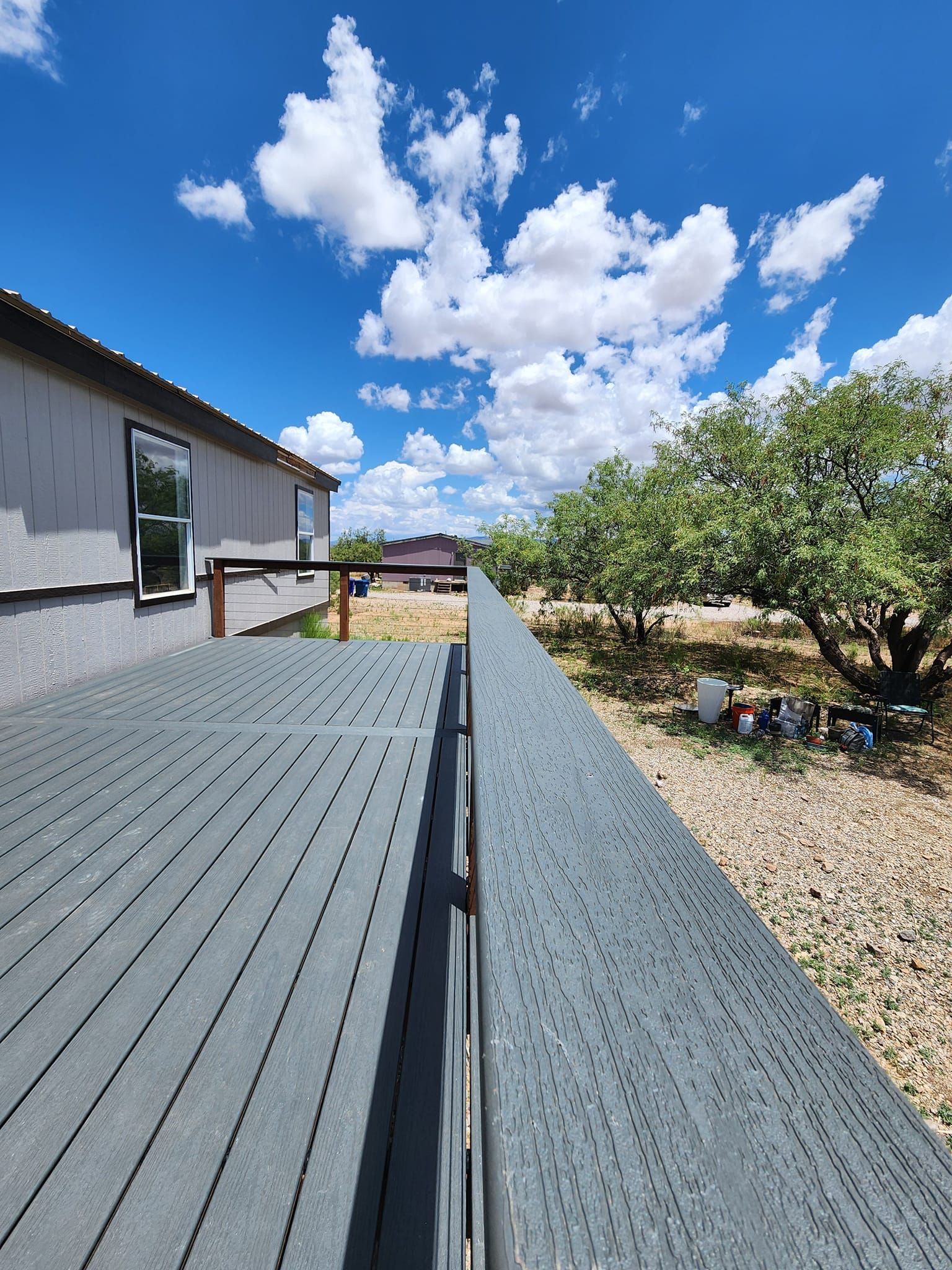 Gray deck with railing, blue sky and clouds. House on the left, tree and gravel yard on the right.