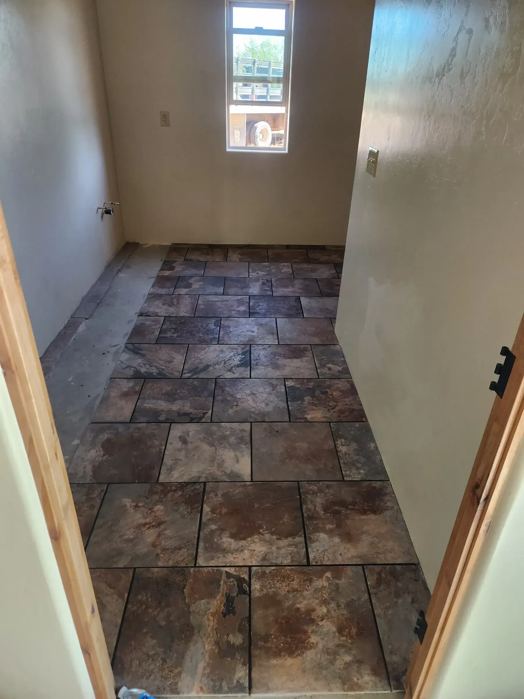 Room with partially laid brown square tiles on the floor, small window, tan walls.