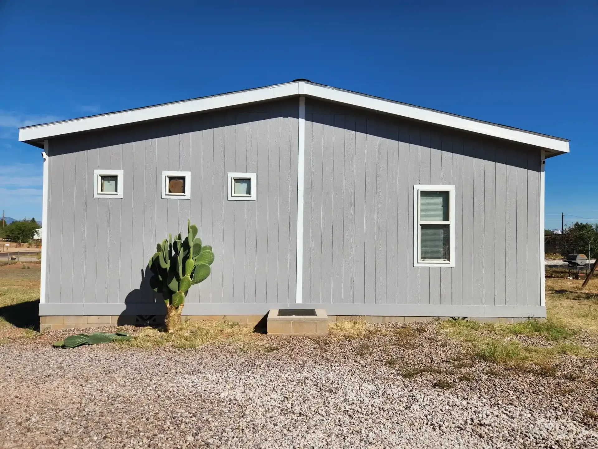 Gray building with white trim, three small windows, one larger window. Cactus in front on gravel.