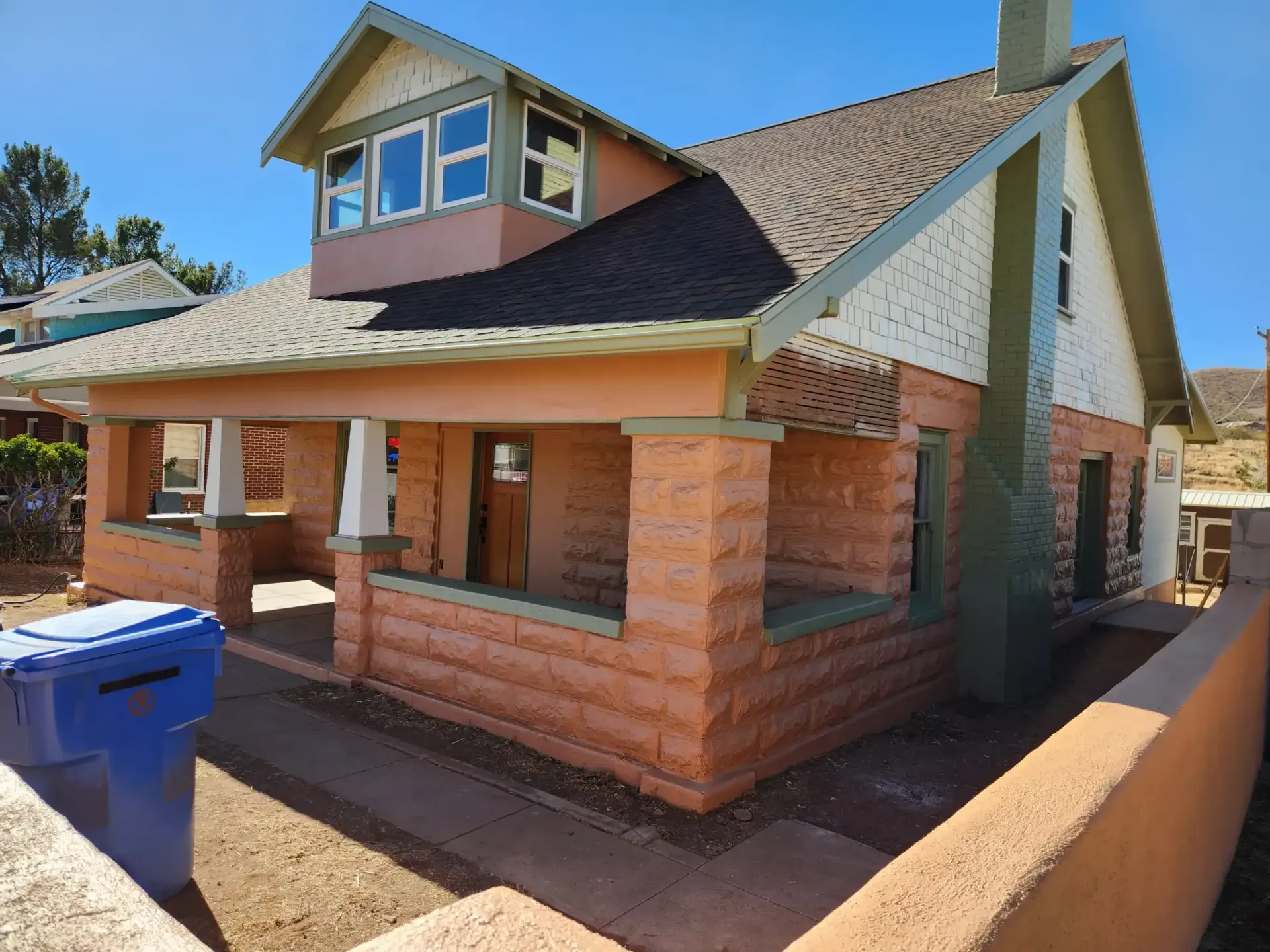 Two-story house with tan brick exterior, porch, and a dormer. Blue trash bin in the foreground.