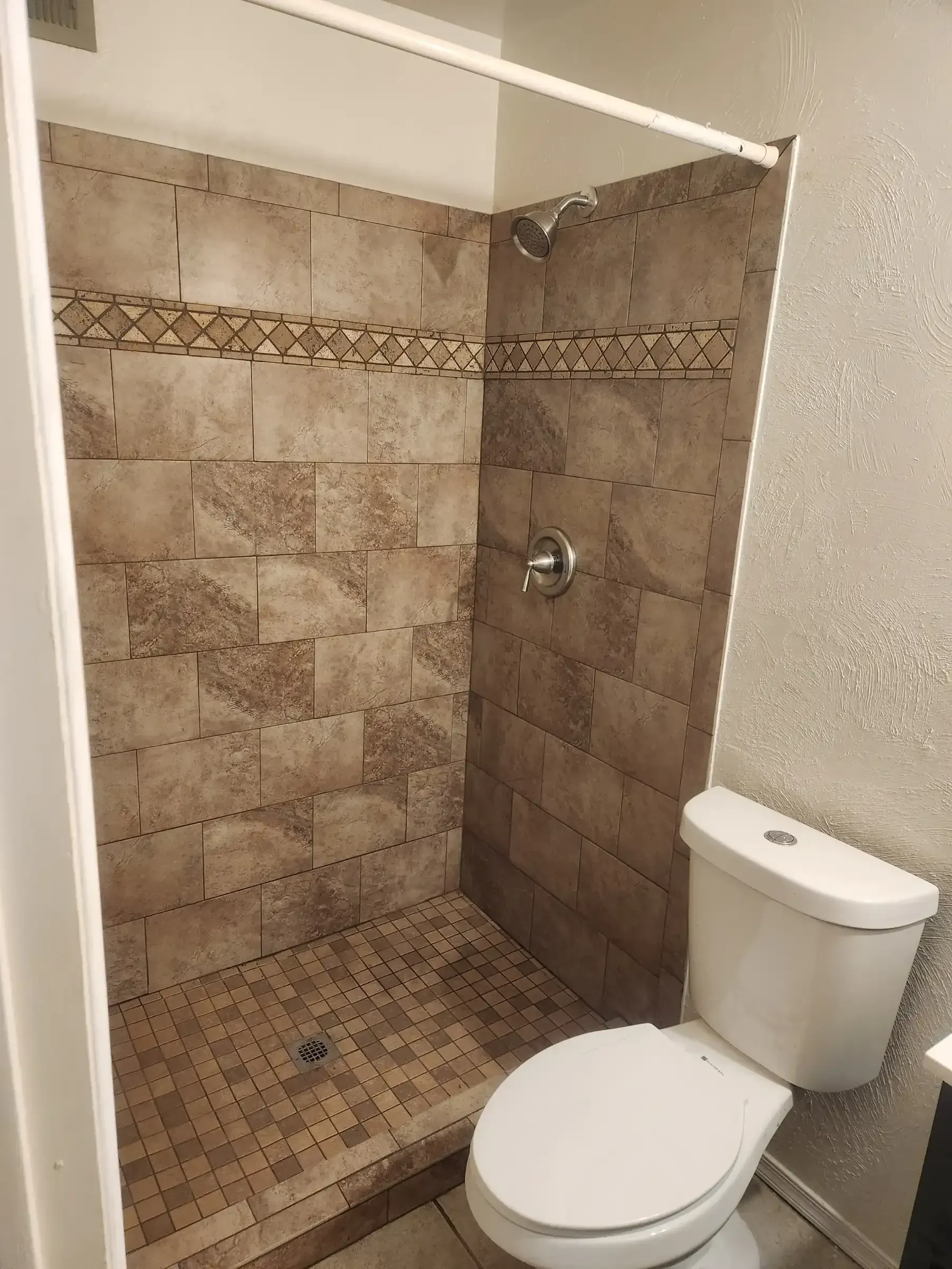 Shower stall with brown tile, toilet on the right. A shower curtain rod is installed above.
