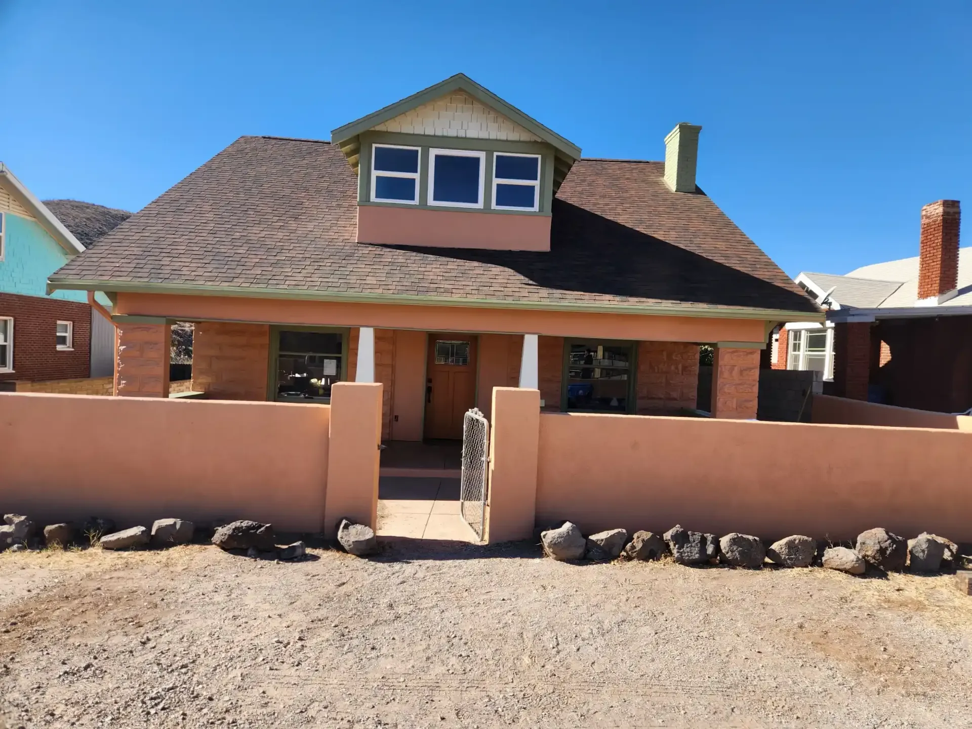 Tan brick house with porch and low wall, brown roof, and blue sky.