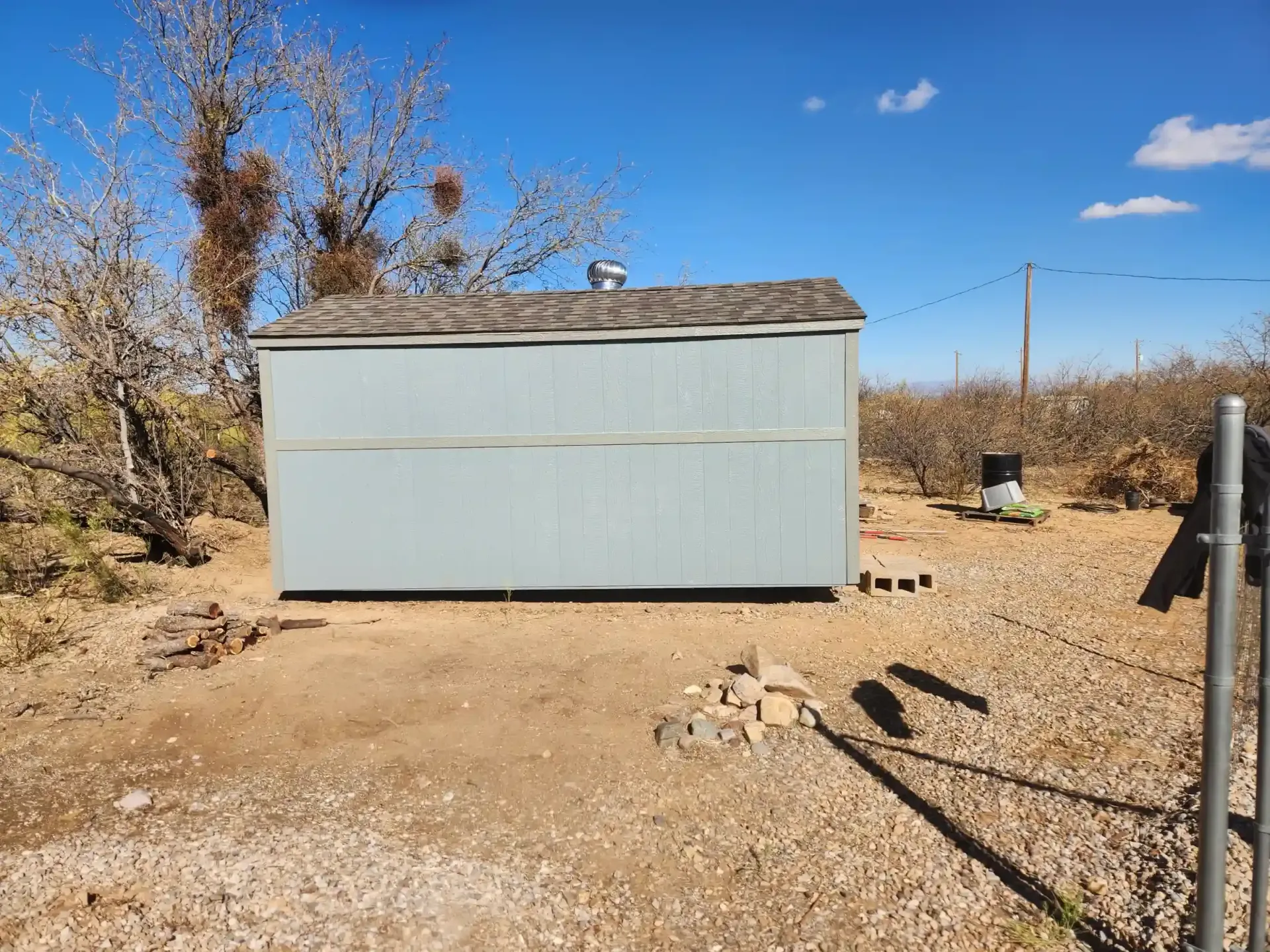 Blue shed in a dirt yard, under a blue sky, surrounded by brown shrubs and trees.