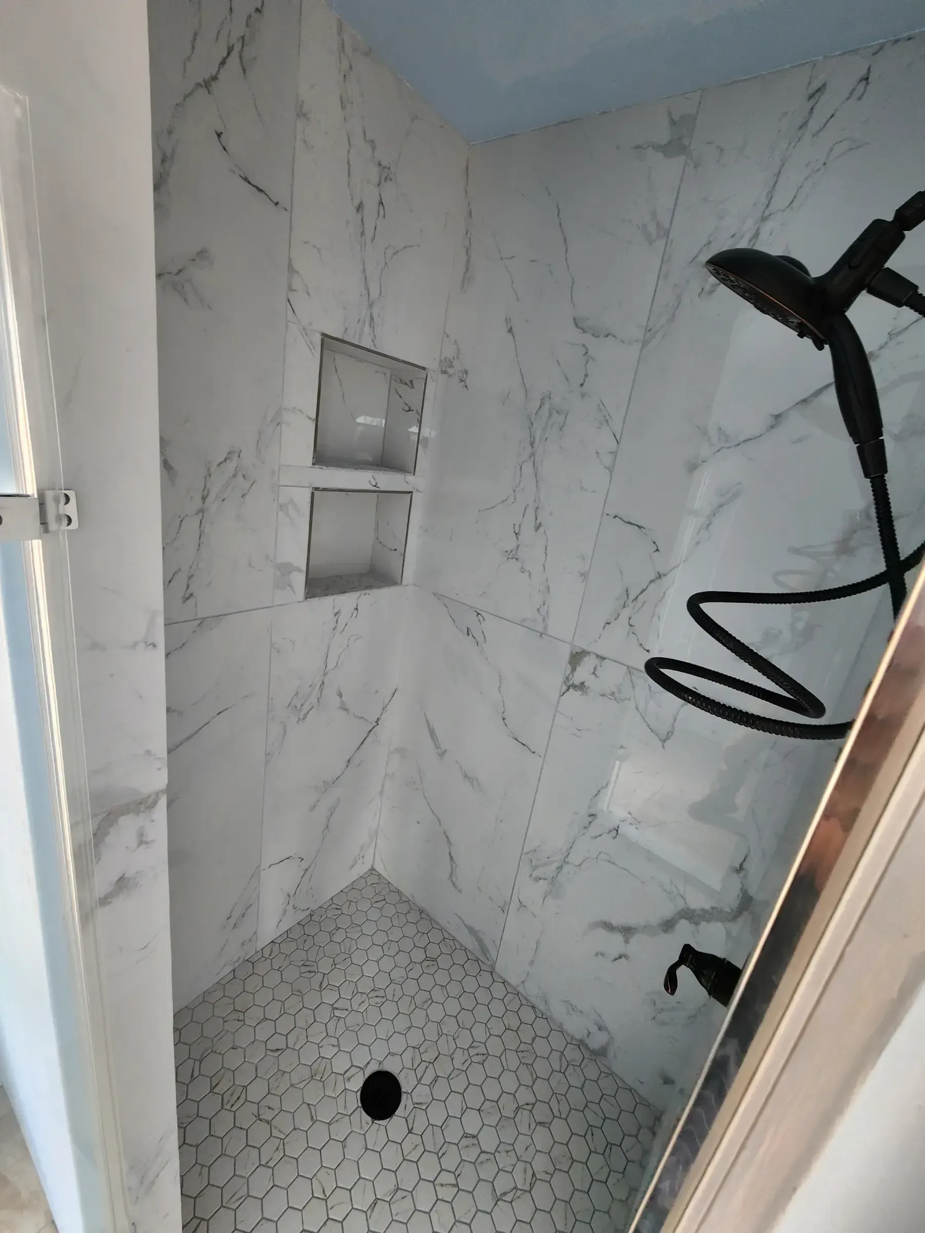 White-tiled shower with a black shower head, pebble floor, and recessed shelf.
