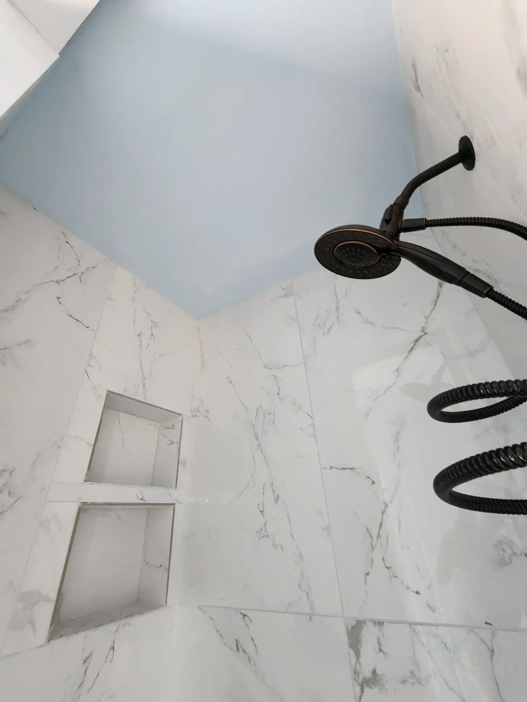 Shower with white marble-look tiles, blue ceiling, and black showerhead with a built-in shelf.