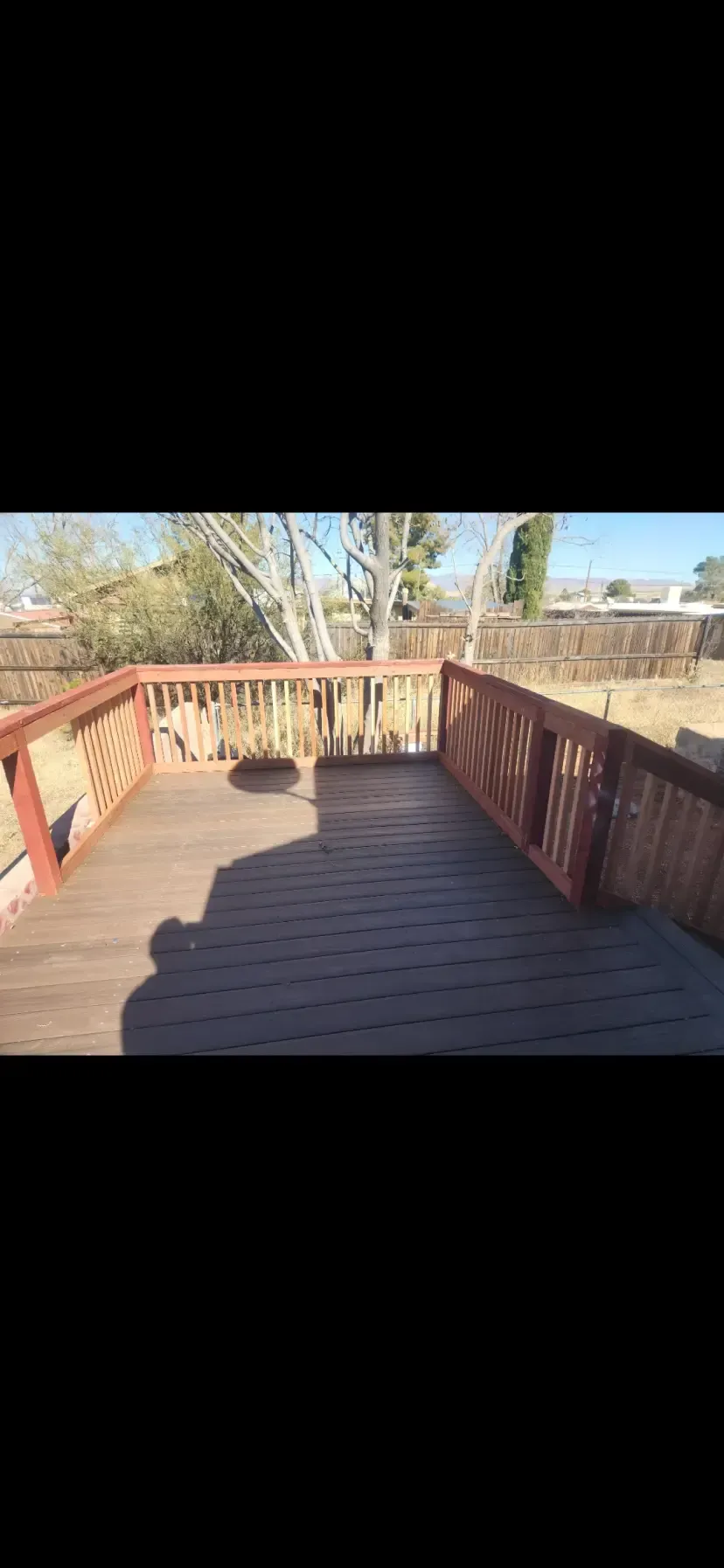 A wooden deck with a brown stain and red railings is in an outdoor setting with a sunny sky.