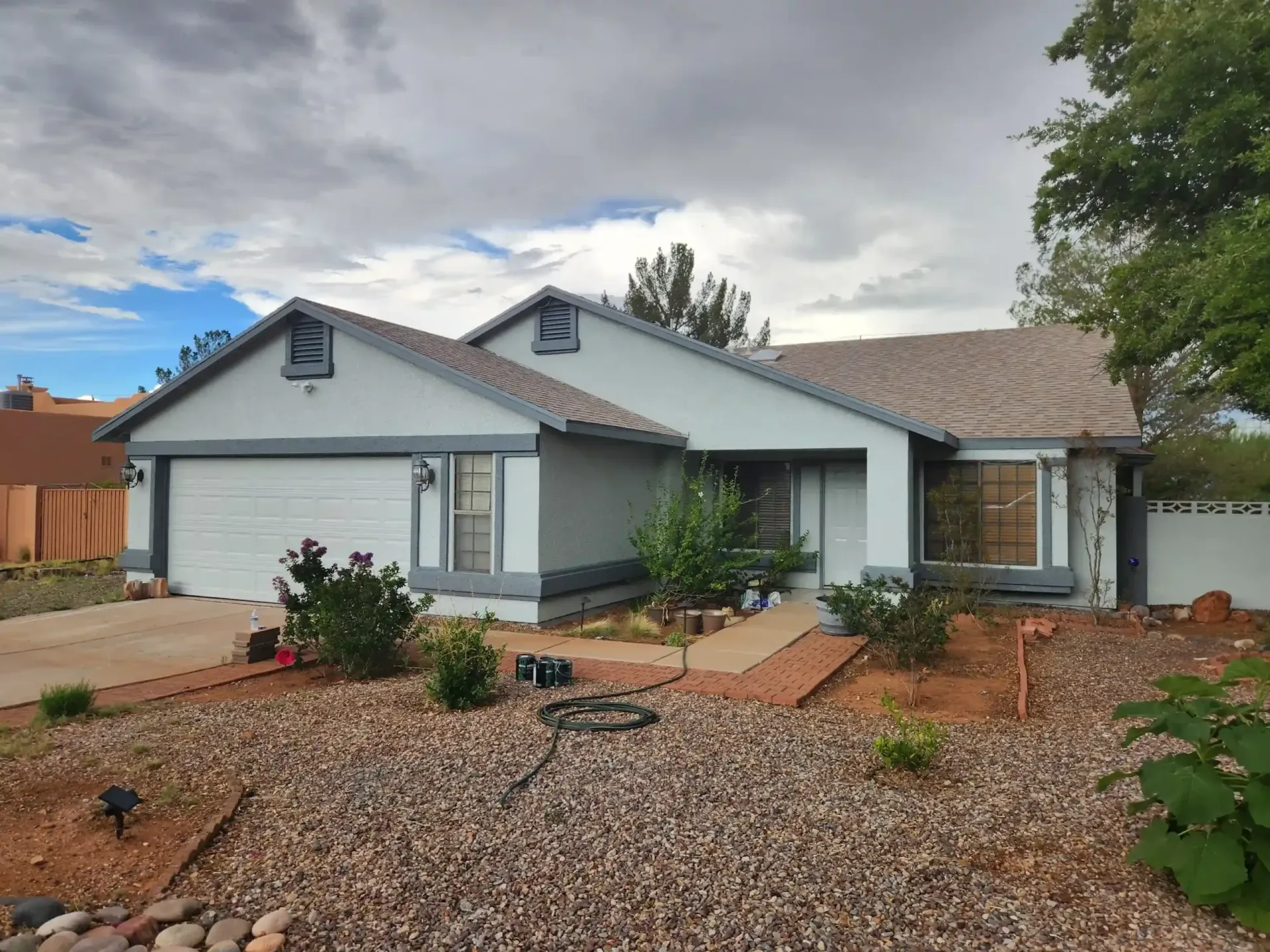 Light blue house with brown roof and gray trim against a cloudy sky. Landscaping with rocks and plants.