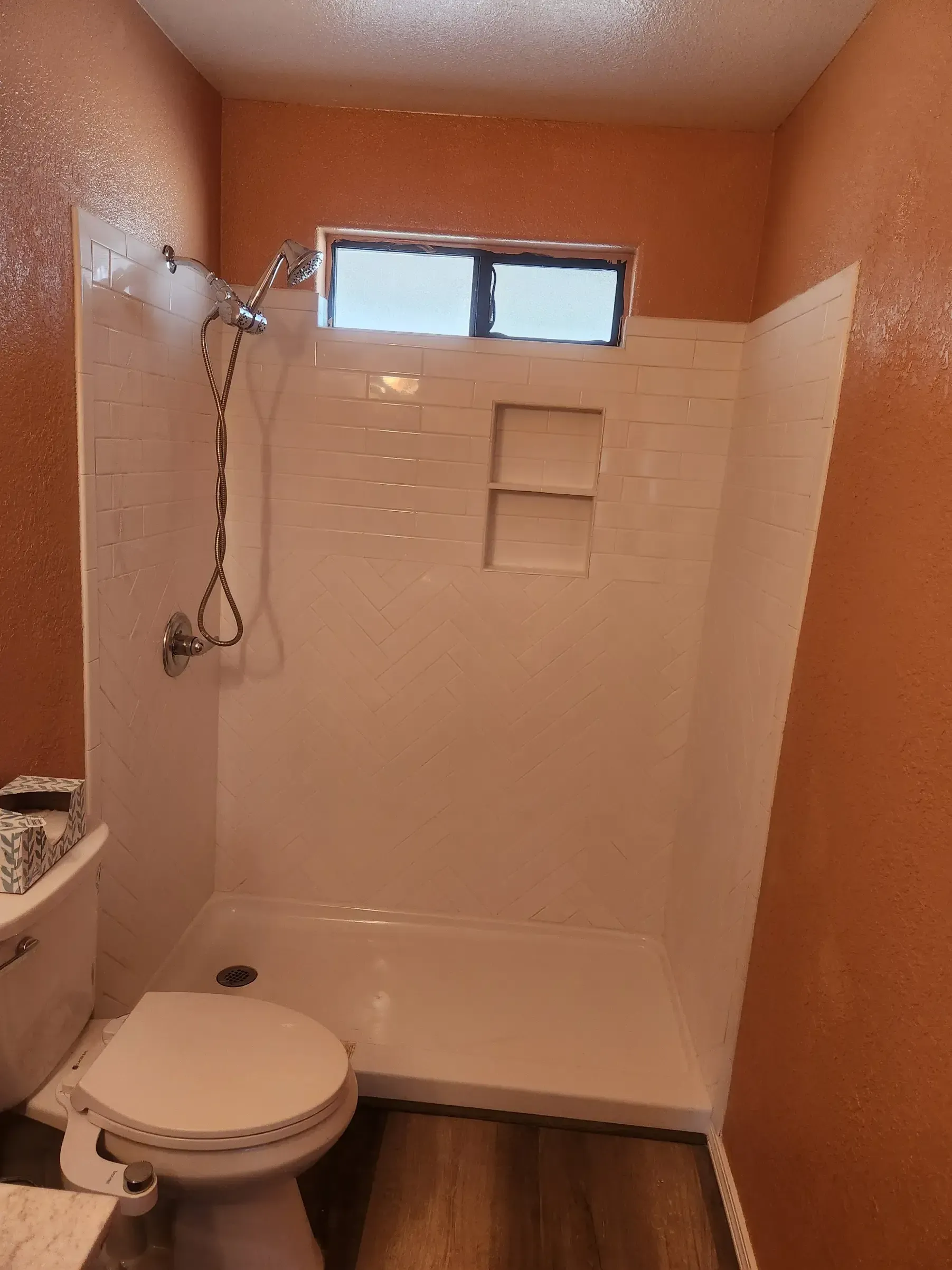 White shower with a built-in shelf in a bathroom with orange walls, toilet visible.