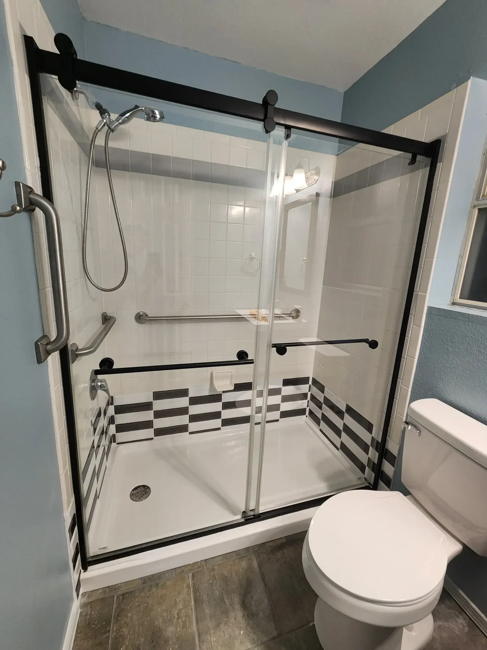 Shower with black sliding glass doors, grab bars, and a toilet. White and blue tiles.
