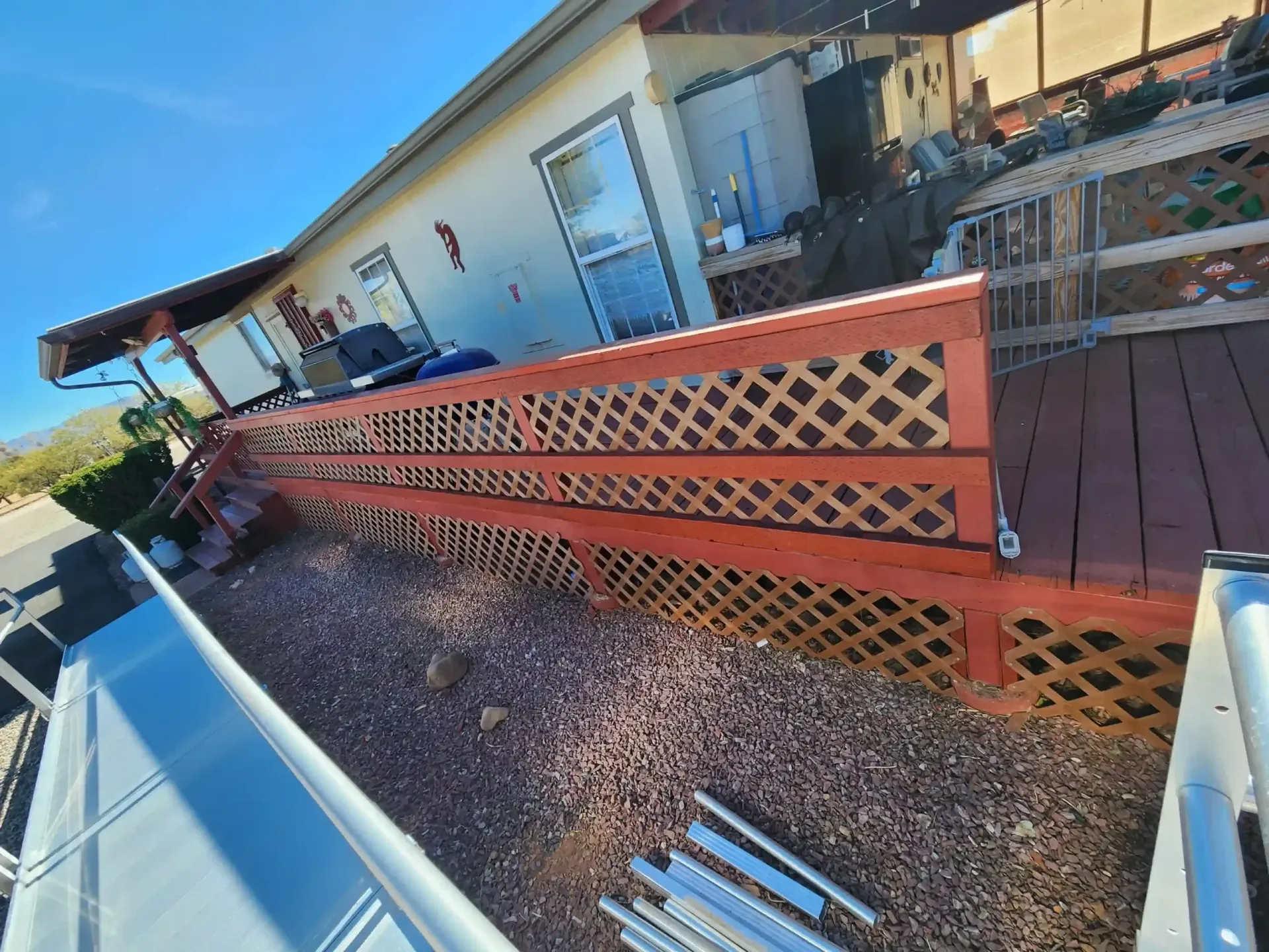 Wooden deck with lattice railing, red-brown paint. House in background, gravel ground.