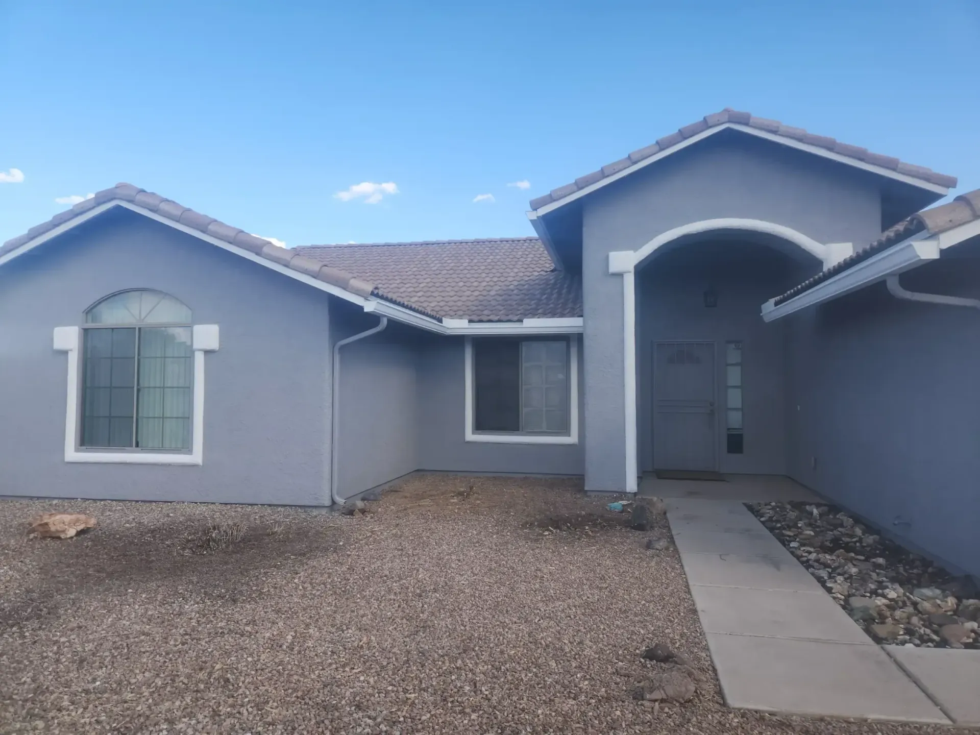Gray house with a tiled roof and white trim. Gravel yard and paved walkway under a blue sky.