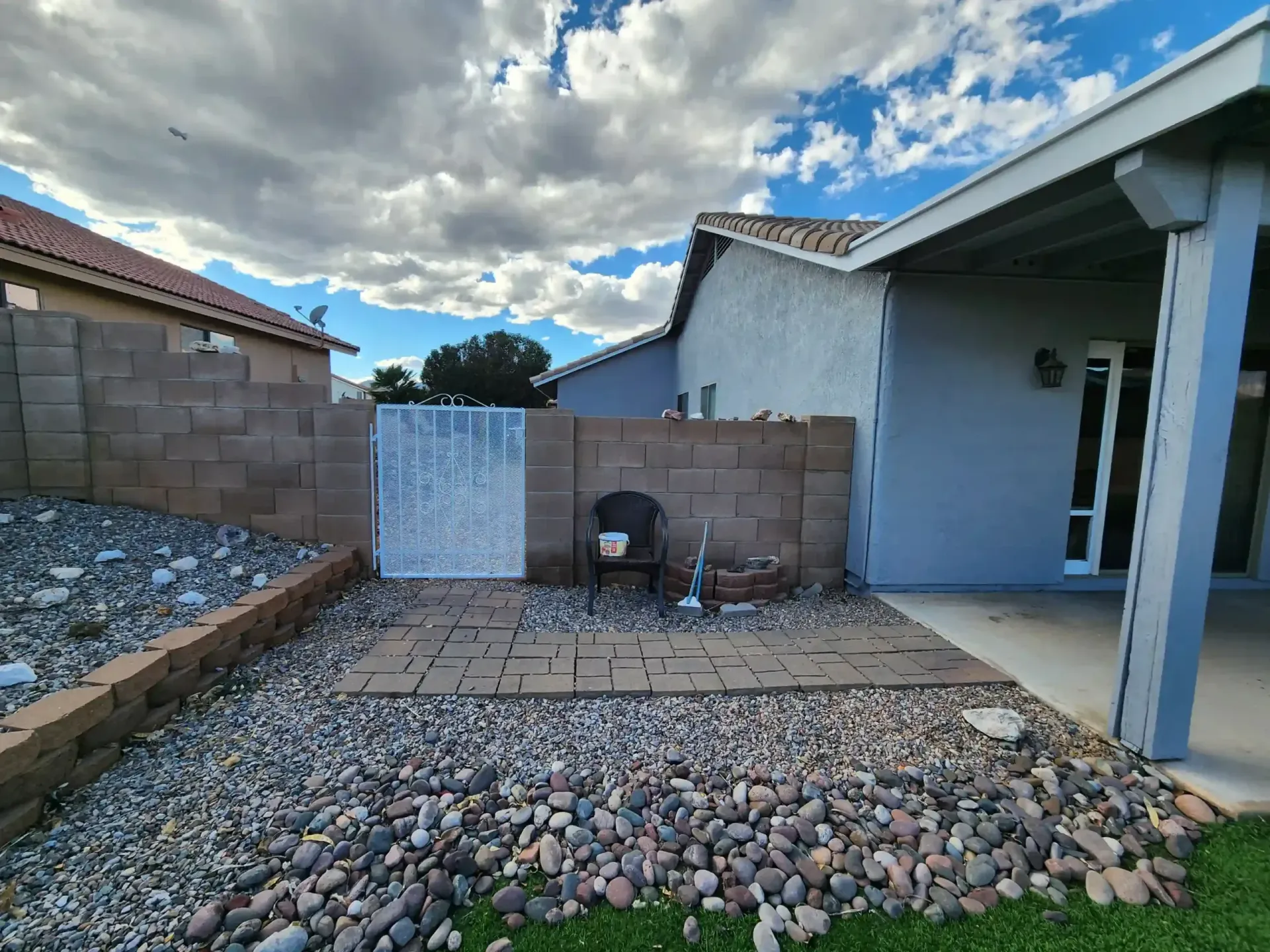 Backyard with stone ground cover, brick walls, a patio, and a blue-gray house under a cloudy sky.