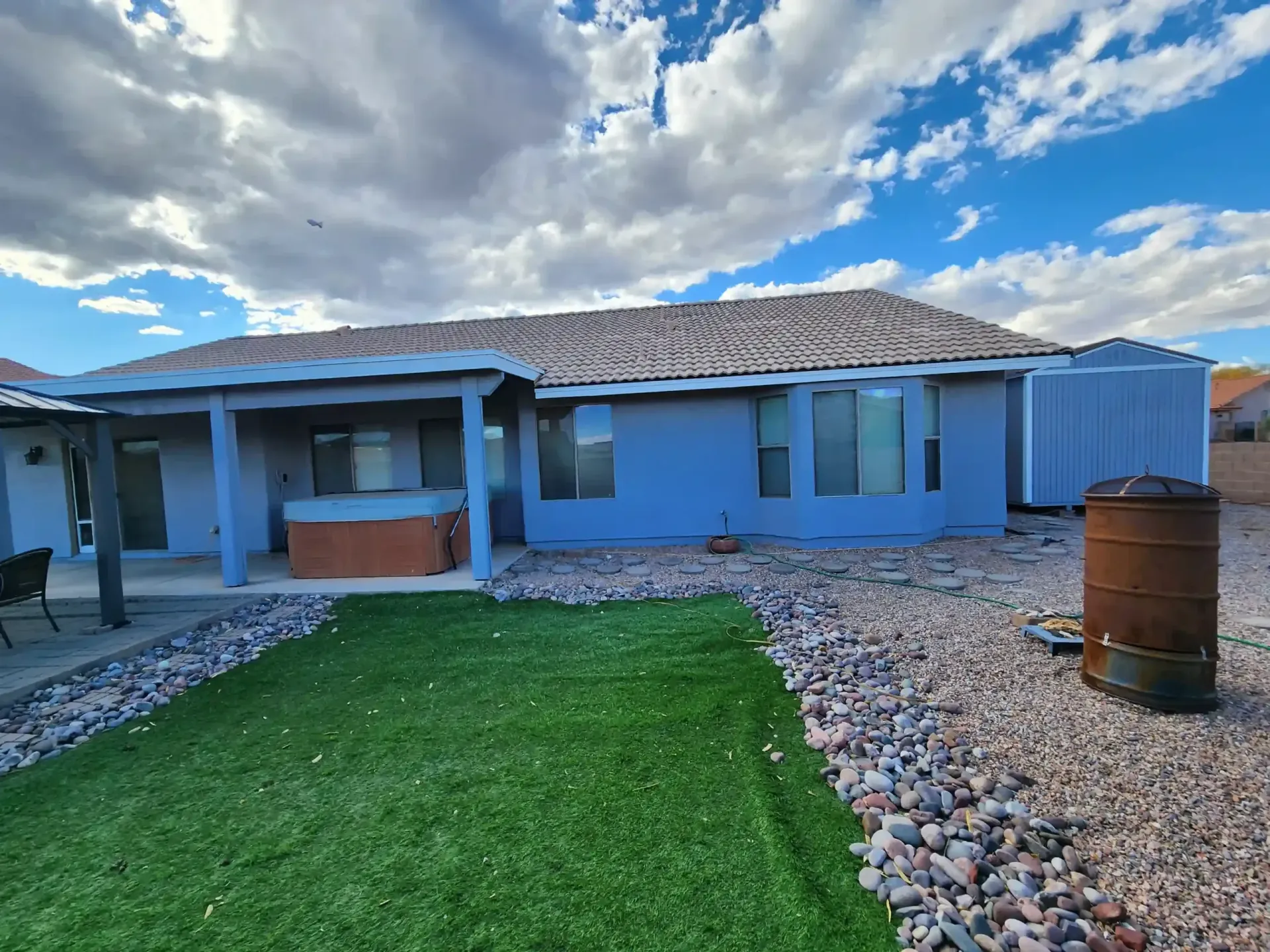 Backyard with a house, a small patch of green turf, gravel, a hot tub, and a blue sky.