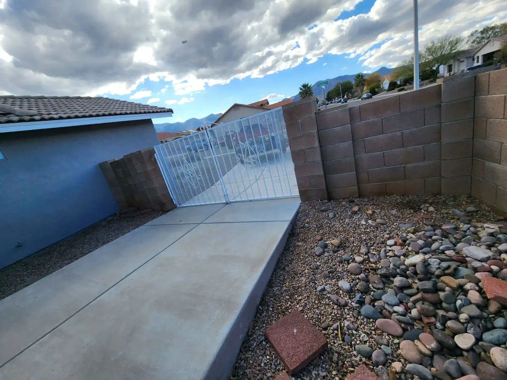 A concrete sidewalk and gravel yard with a leaning wall against a house under a cloudy sky.