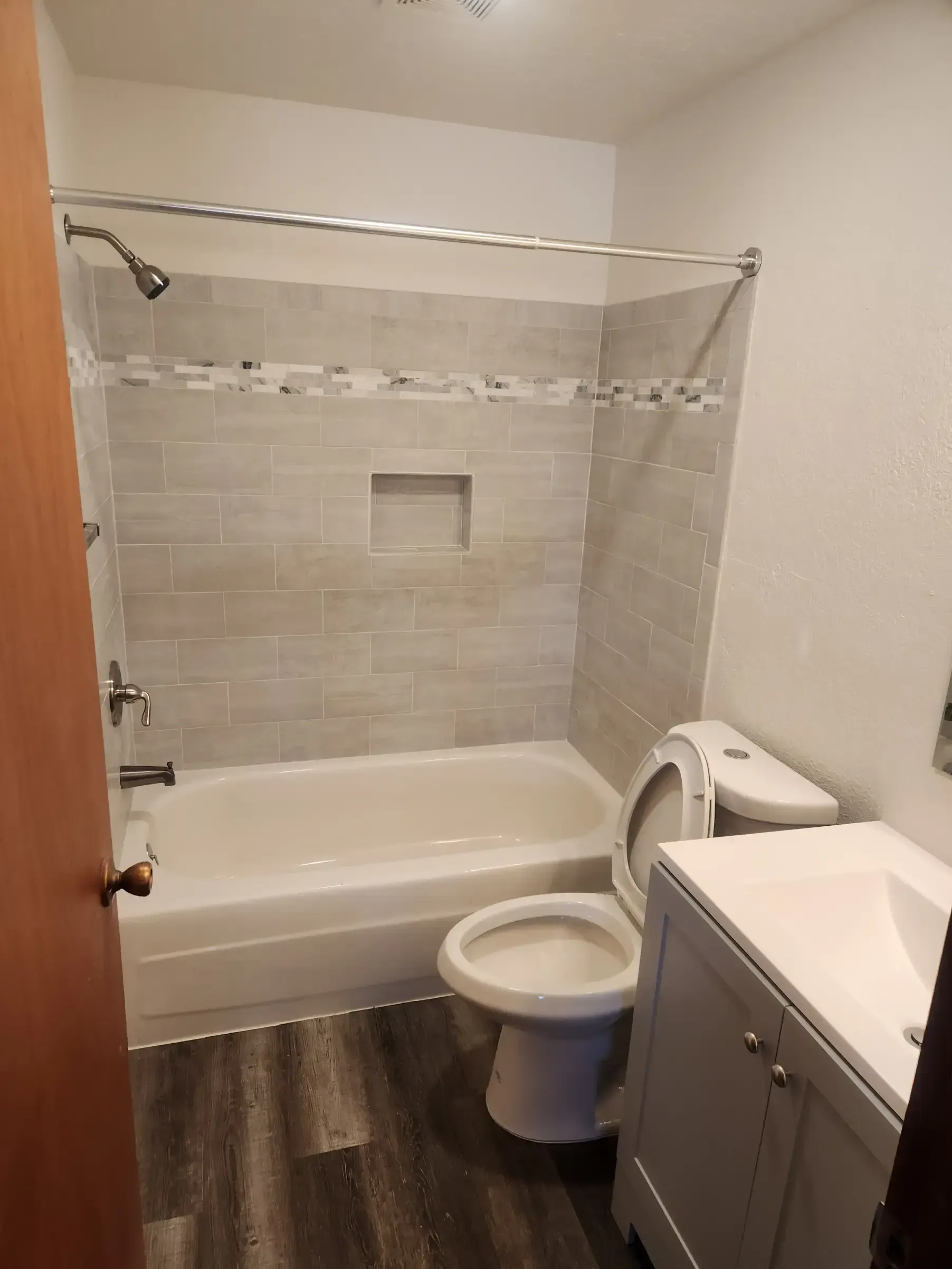 Bathroom with a bathtub, toilet, and vanity. Gray tile surround and wood-look flooring.
