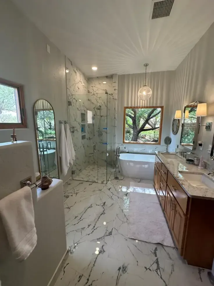 Modern bathroom with marble floors, glass shower, freestanding tub, and a large window overlooking trees.