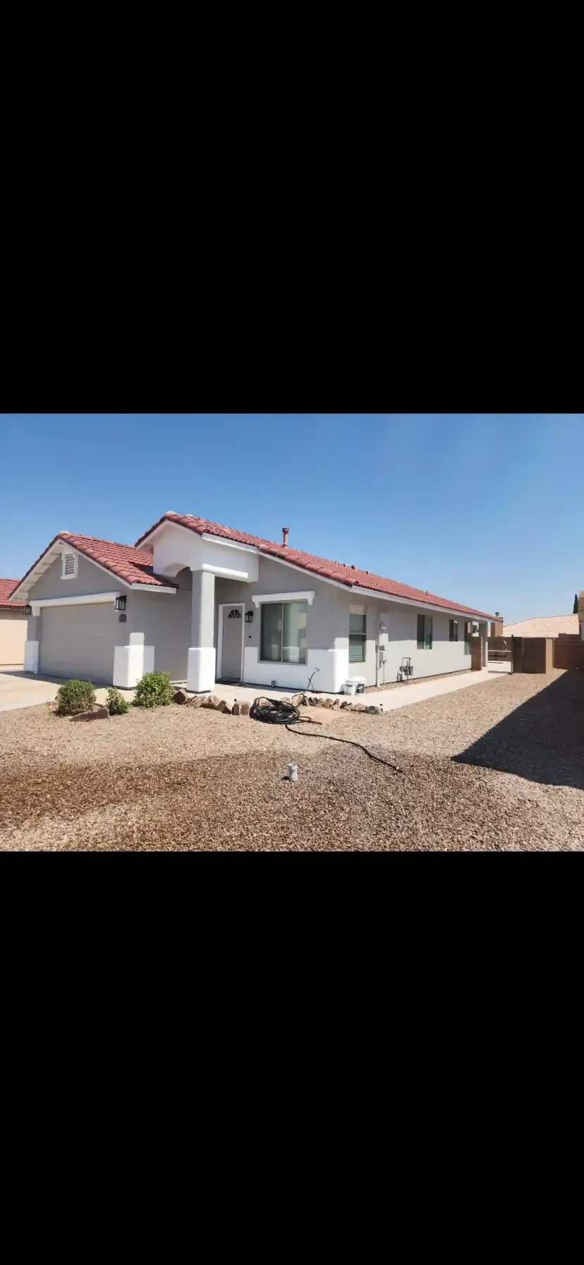 Gray house with red-tiled roof, white pillars, and garage on a gravel lot under a blue sky.