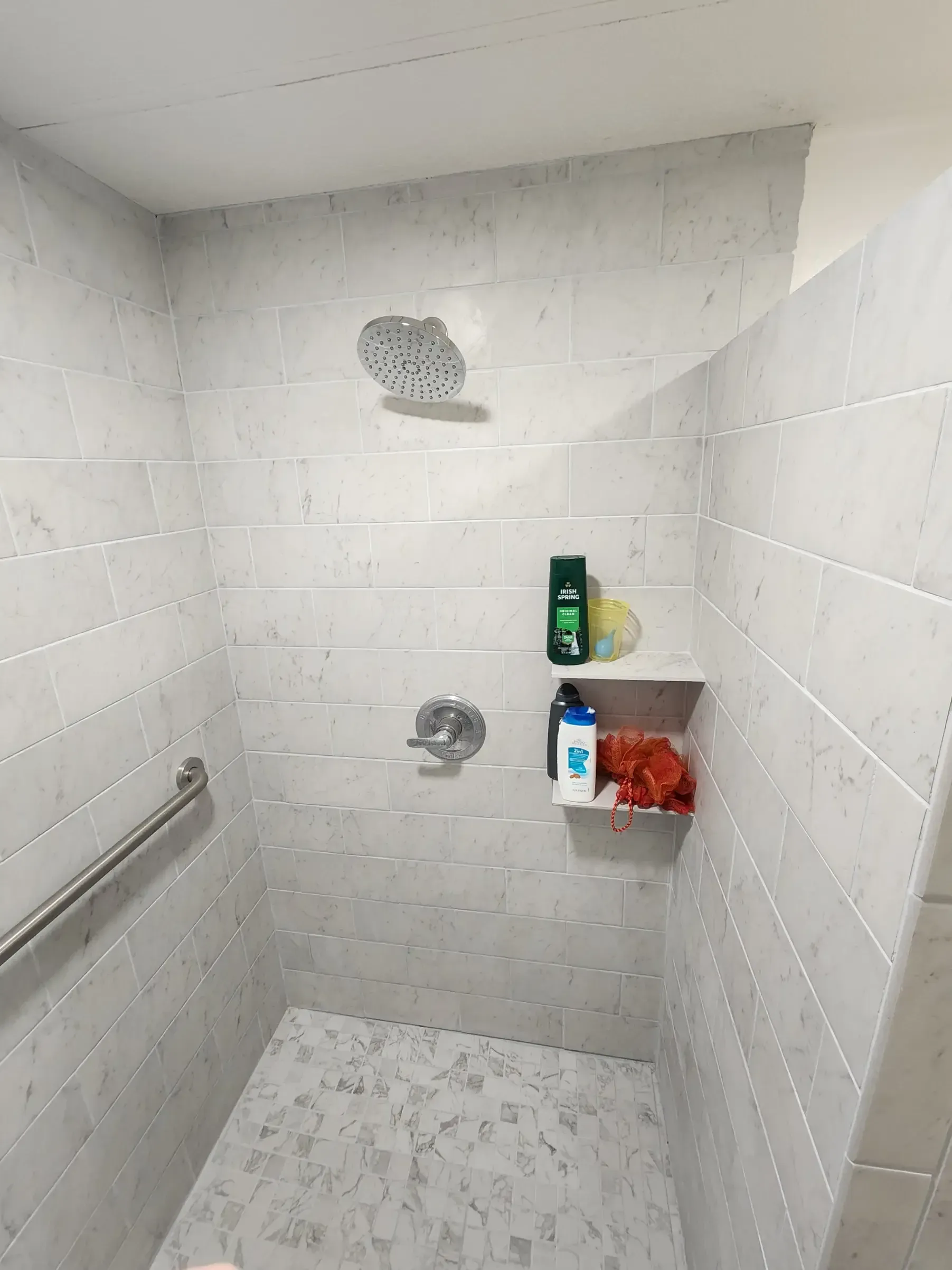 Accessible shower stall with white and gray tile, showerhead, shelf with toiletries, and a grab bar.