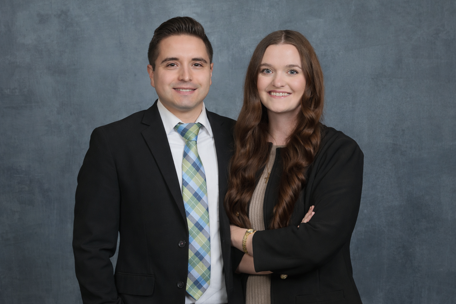Man in suit with arm around woman; both smiling, standing in front of a neutral backdrop.