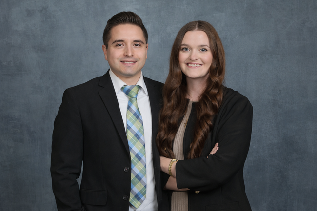 Man in suit with arm around woman; both smiling, standing in front of a neutral backdrop.