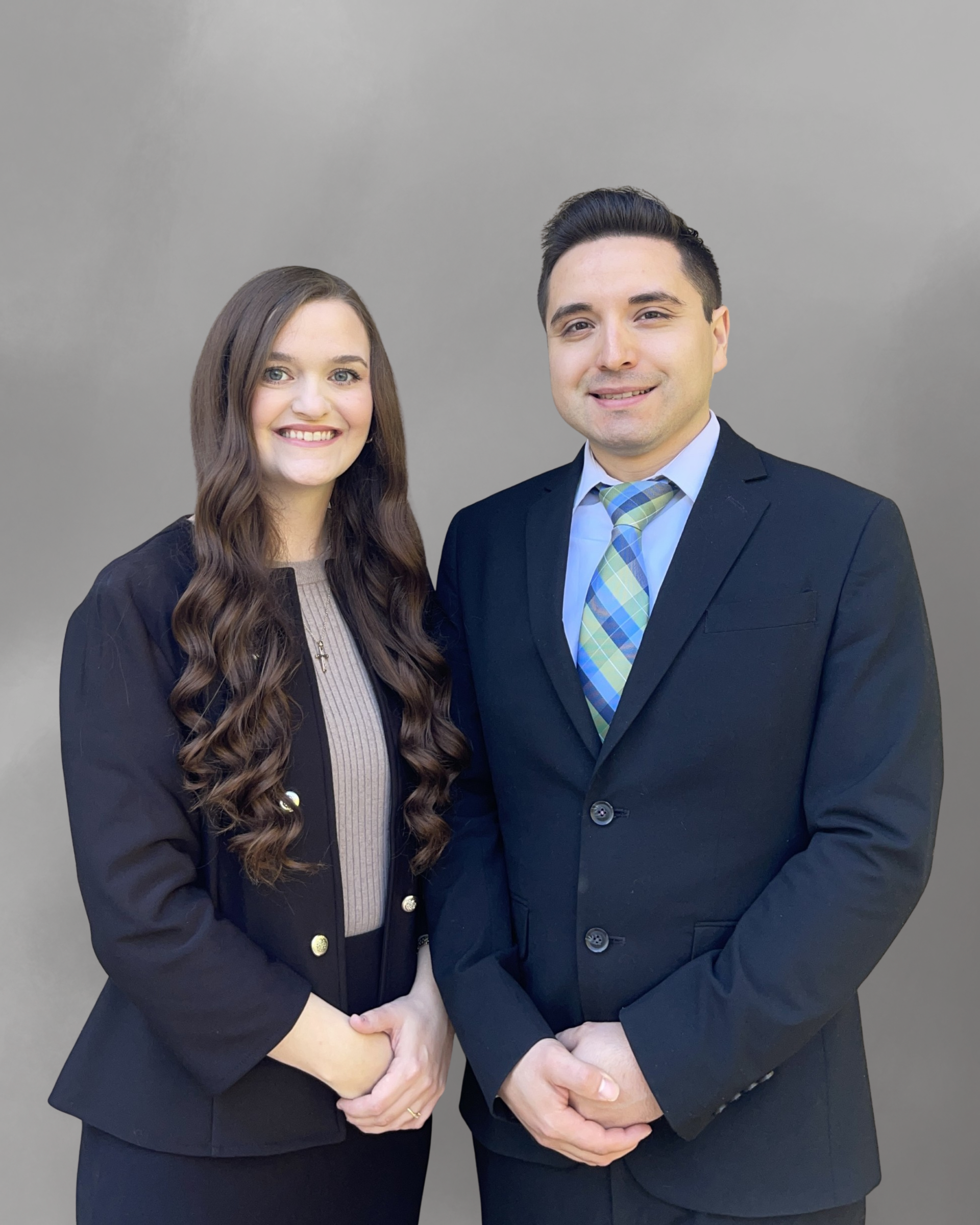 Two people in business attire standing close together, smiling. Gray background.