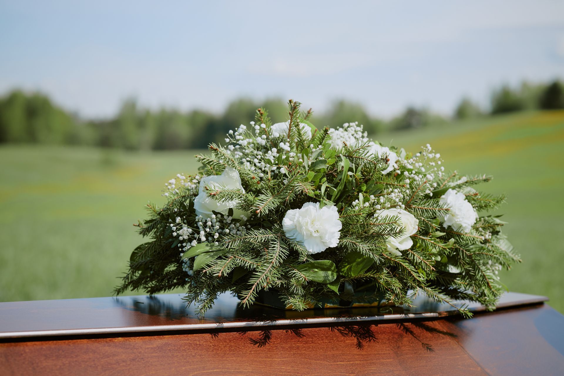 White floral arrangement on a wooden surface, with a green field and trees in the background.