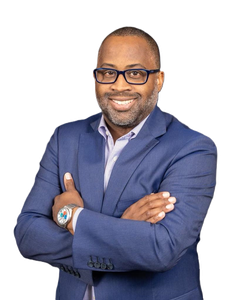 A man in a blue suit and glasses is standing with his arms crossed and smiling.