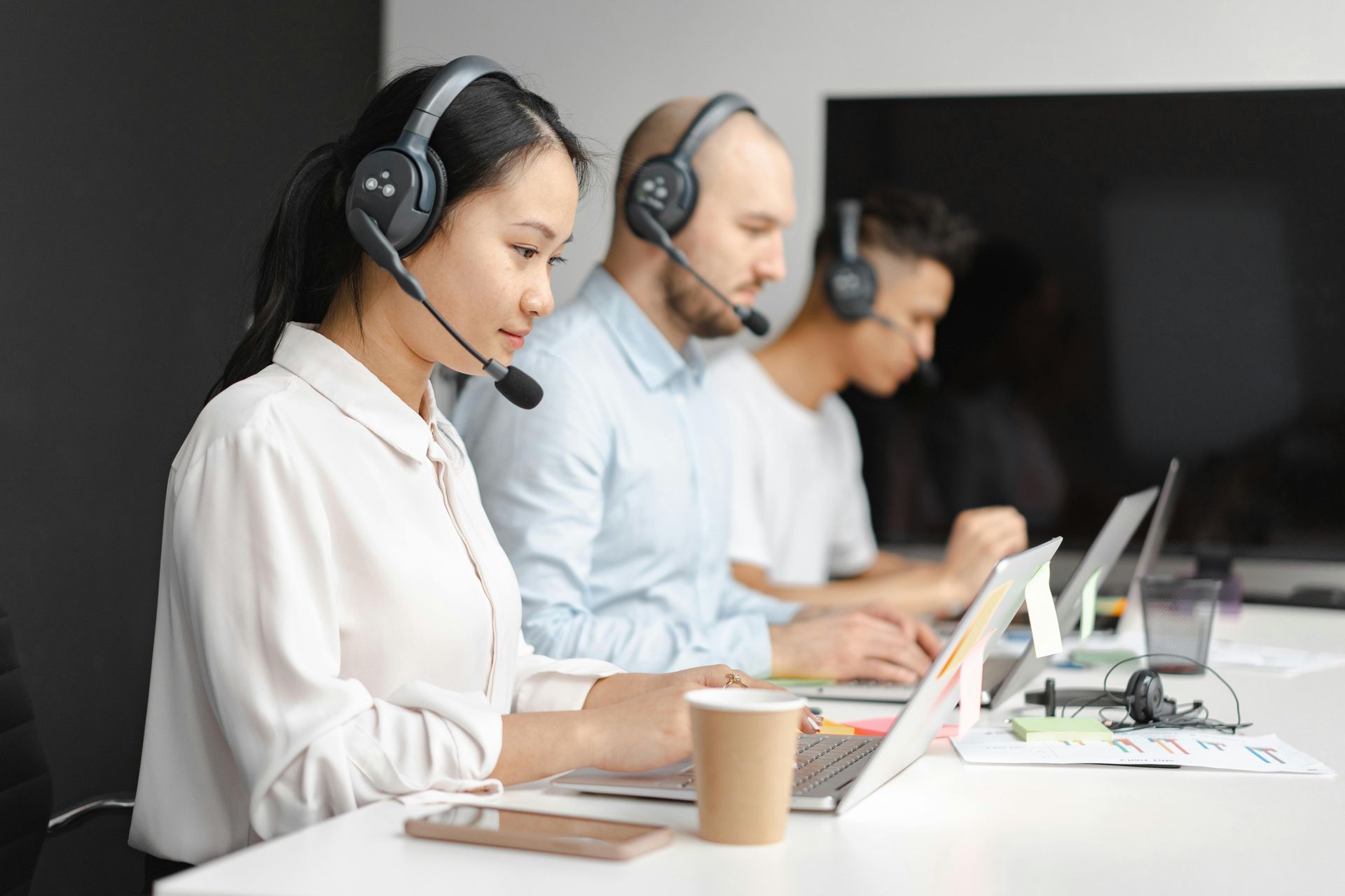 Three people in a modern office, wearing headsets, typing on laptops. They are focused, working at desks.