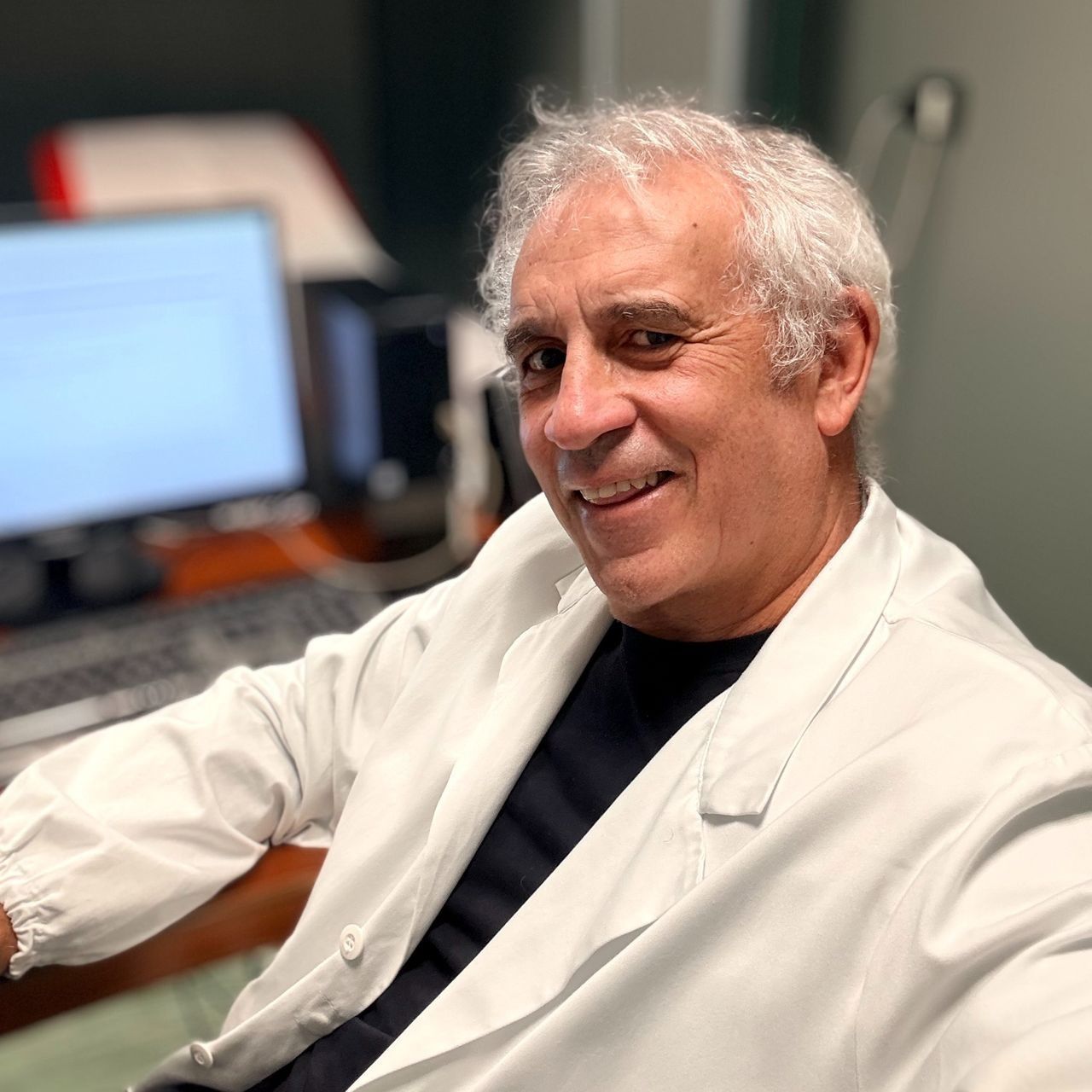 Una persona con i capelli grigi, che indossa un camice bianco, è seduta in un ufficio con un computer sullo sfondo.