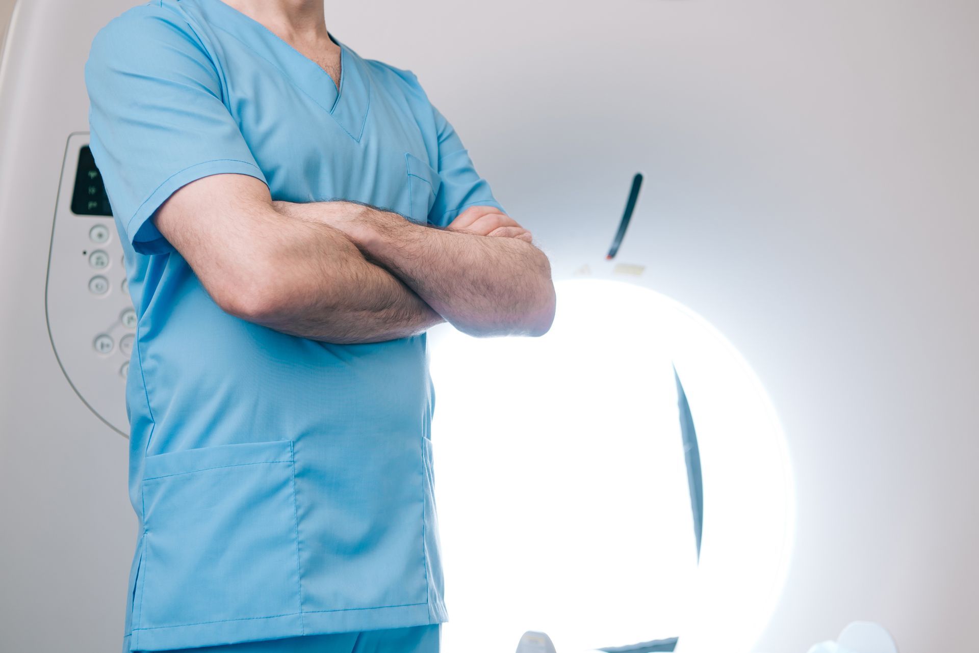 Un operatore sanitario in camice blu è in piedi con le braccia incrociate davanti a un apparecchio per la diagnostica per immagini.
