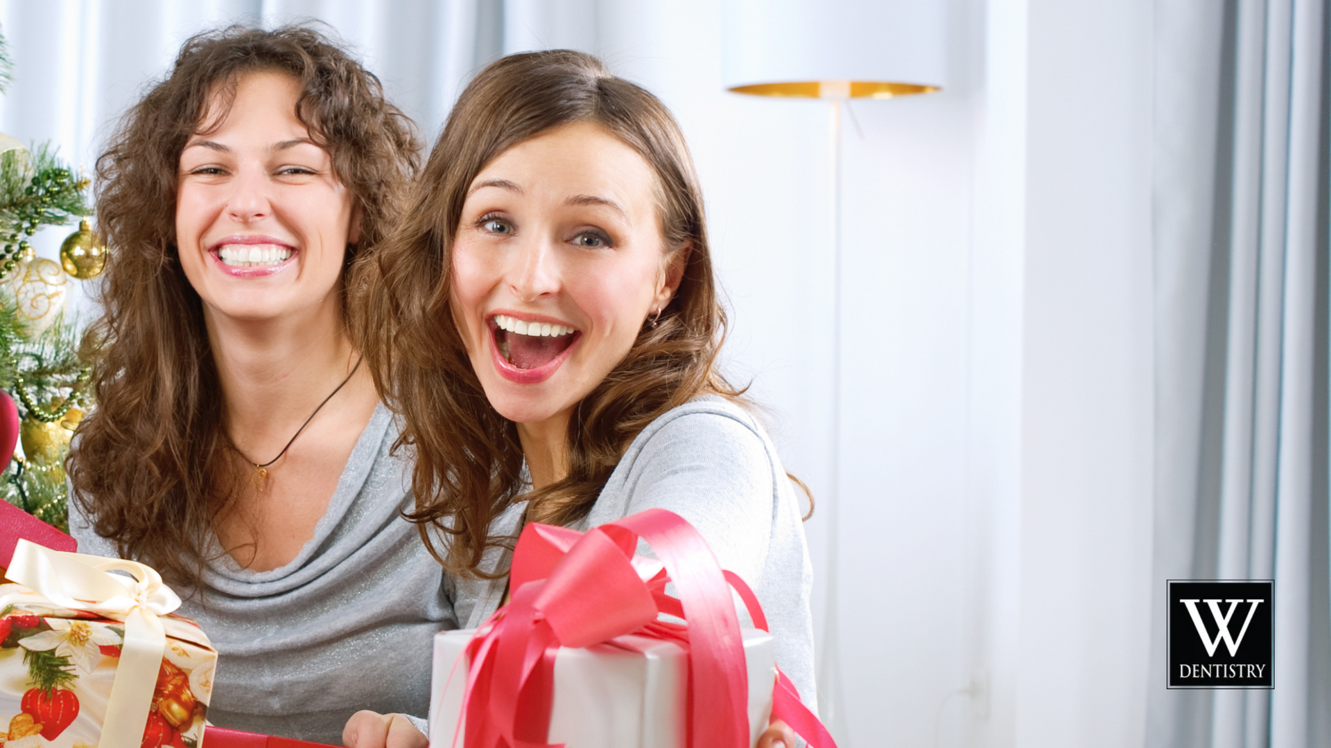 Two women are holding christmas presents in front of a christmas tree.