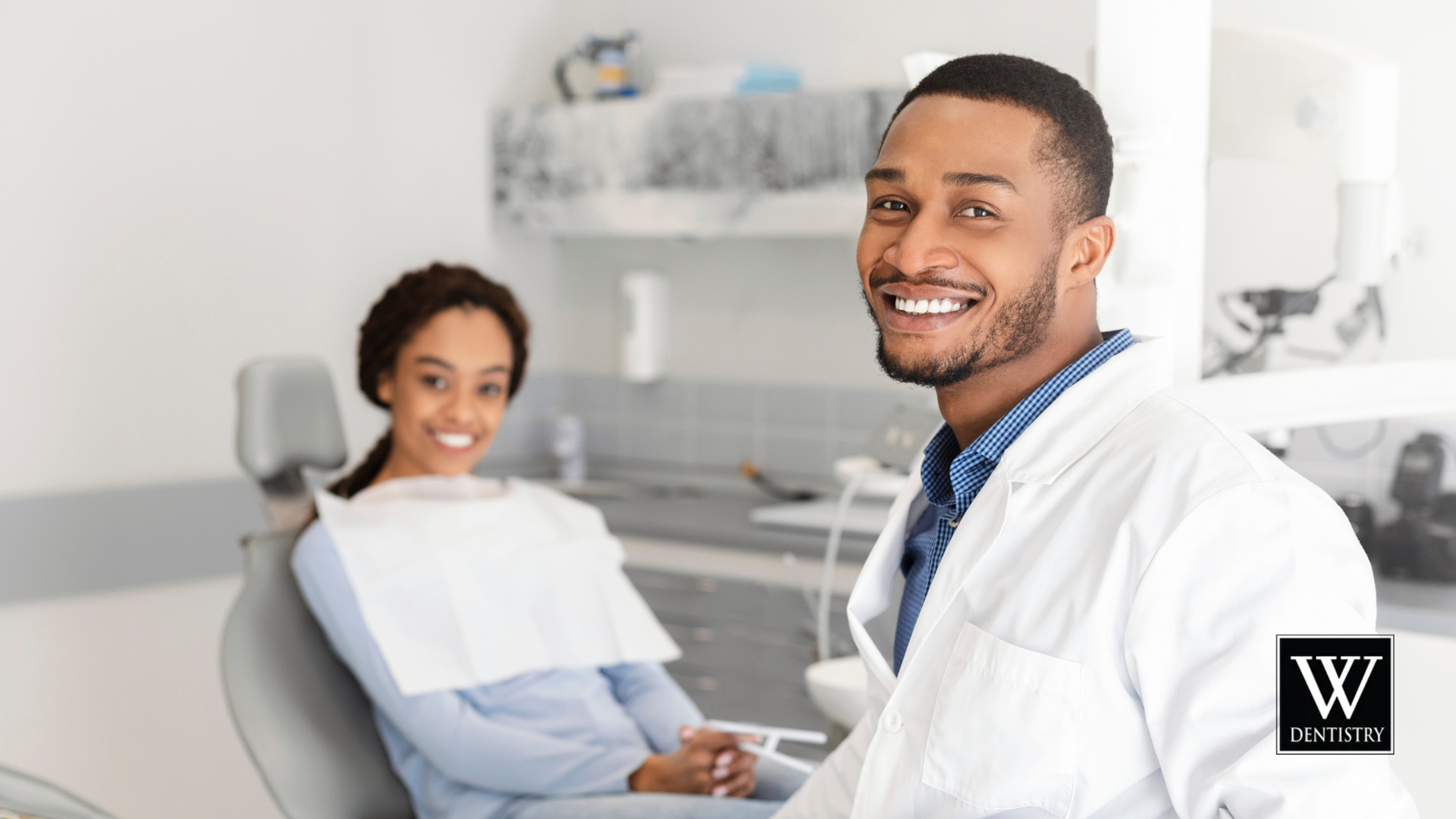 A dentist is smiling while a patient sits in a dental chair.