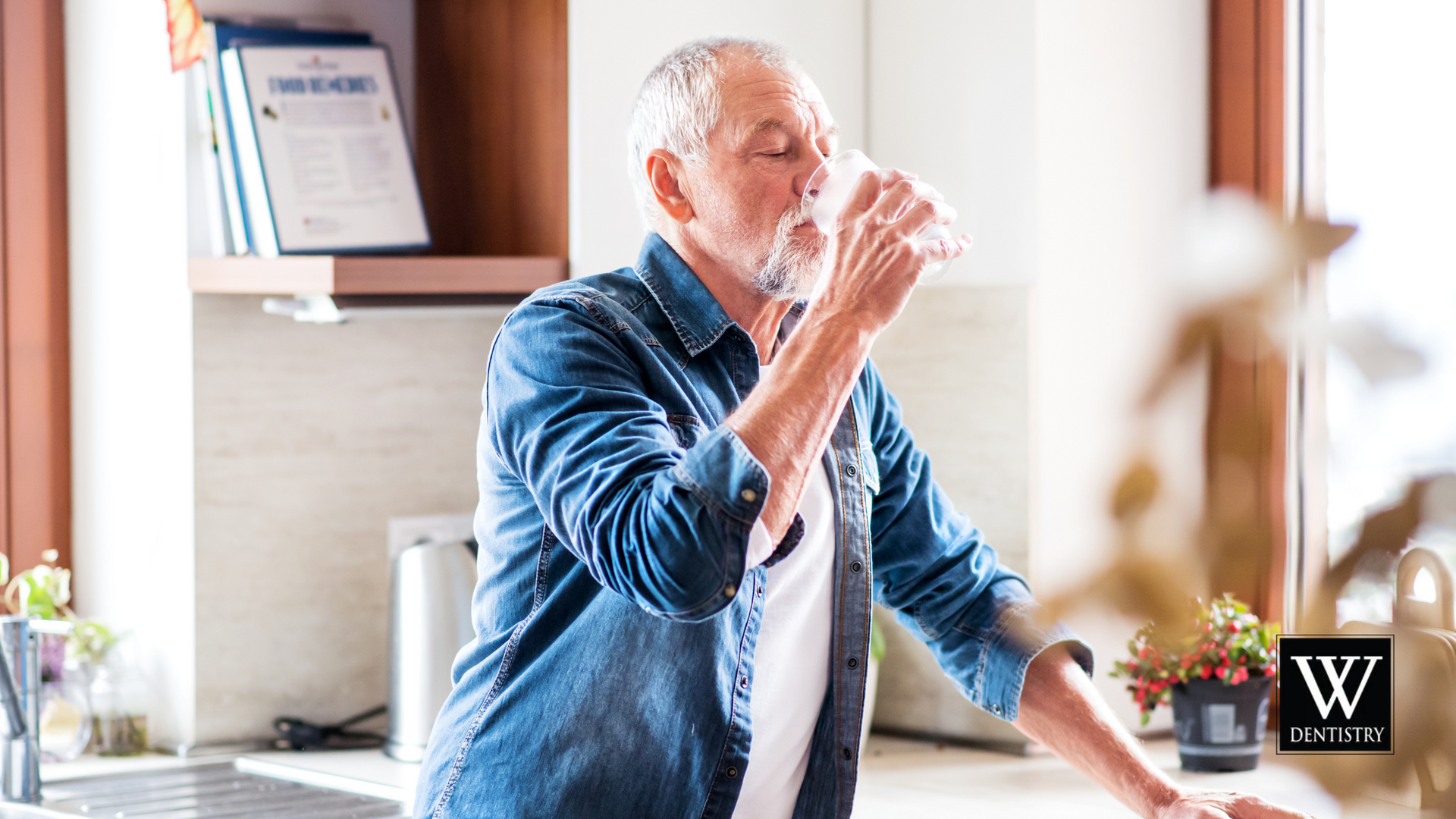 An older man is drinking a glass of water in a kitchen.