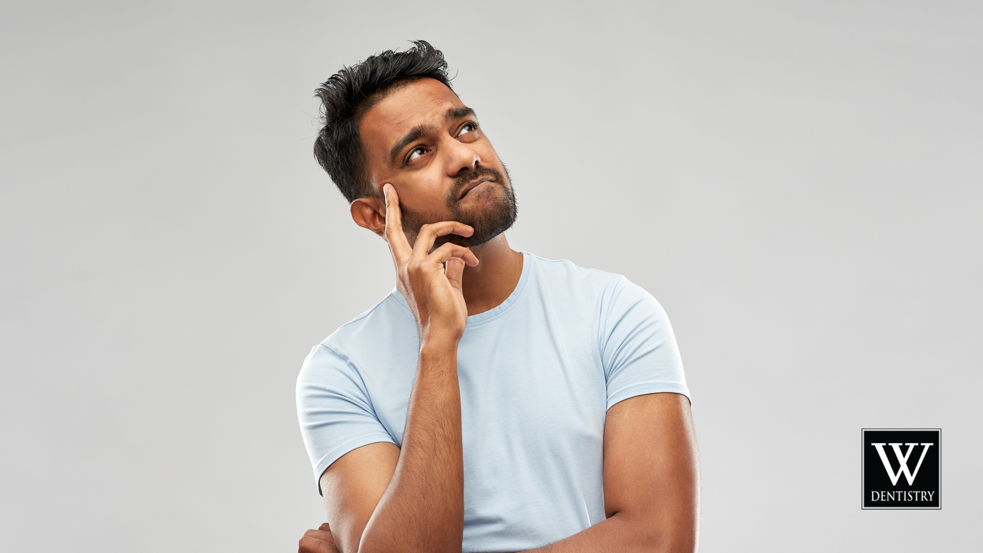 Man in blue shirt, hand on chin, looking upwards thoughtfully. Gray background.