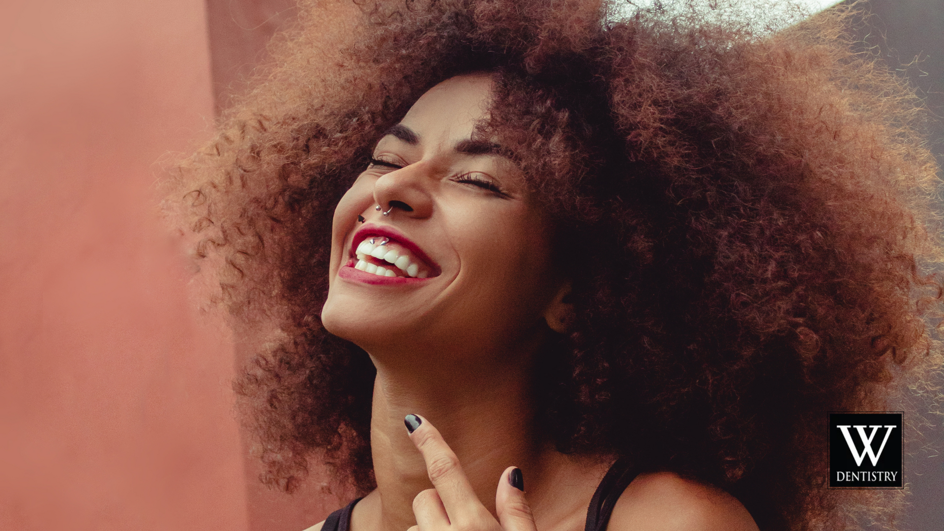 A woman with curly hair is smiling and pointing at her face.