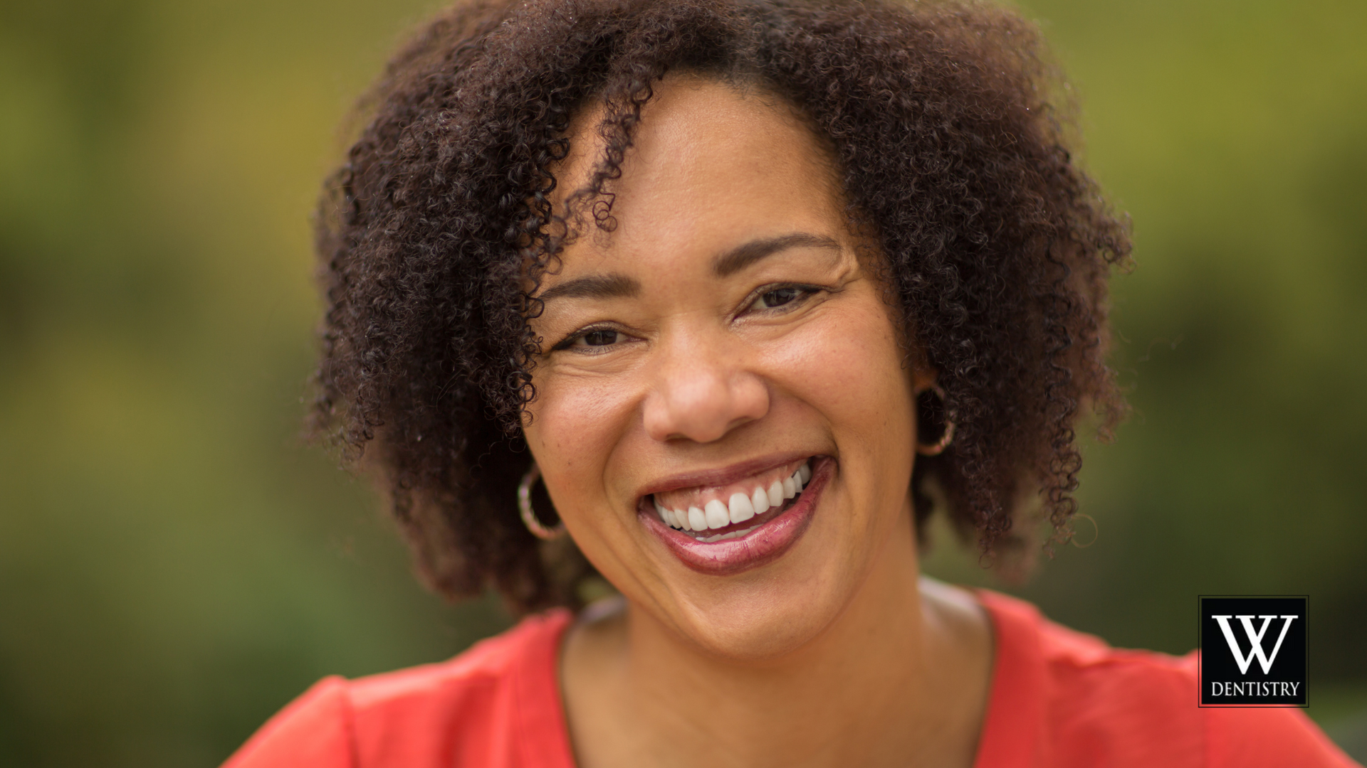 Smiling Black woman with curly hair, wearing red shirt, outdoors.