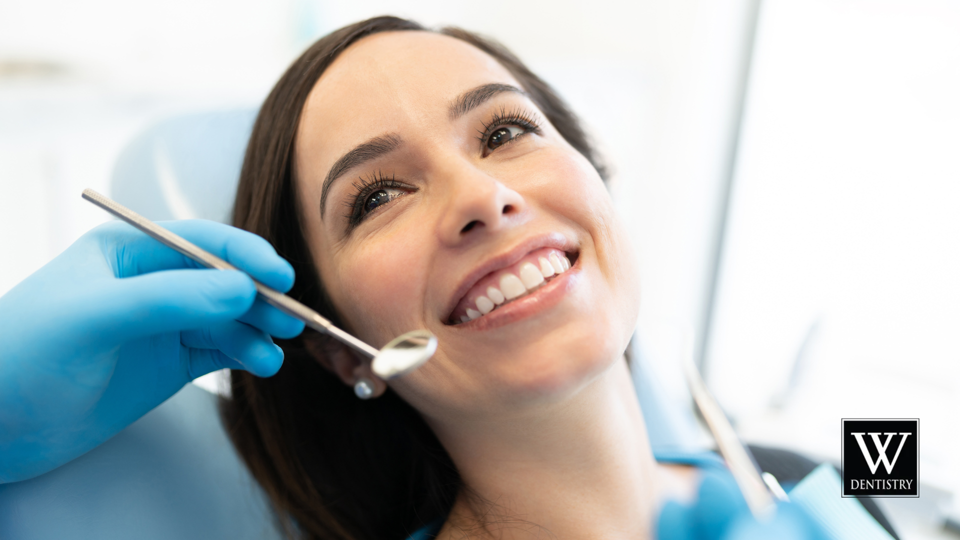 Woman smiling at the dentist during an examination. The dentist holds tools, and is wearing gloves.