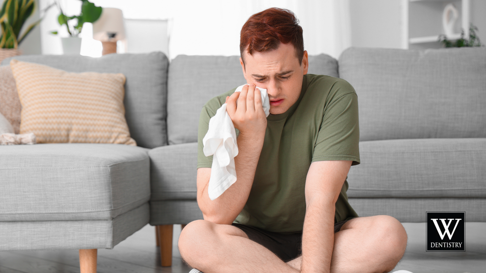 A man is sitting on the floor in front of a couch holding a napkin to his face.