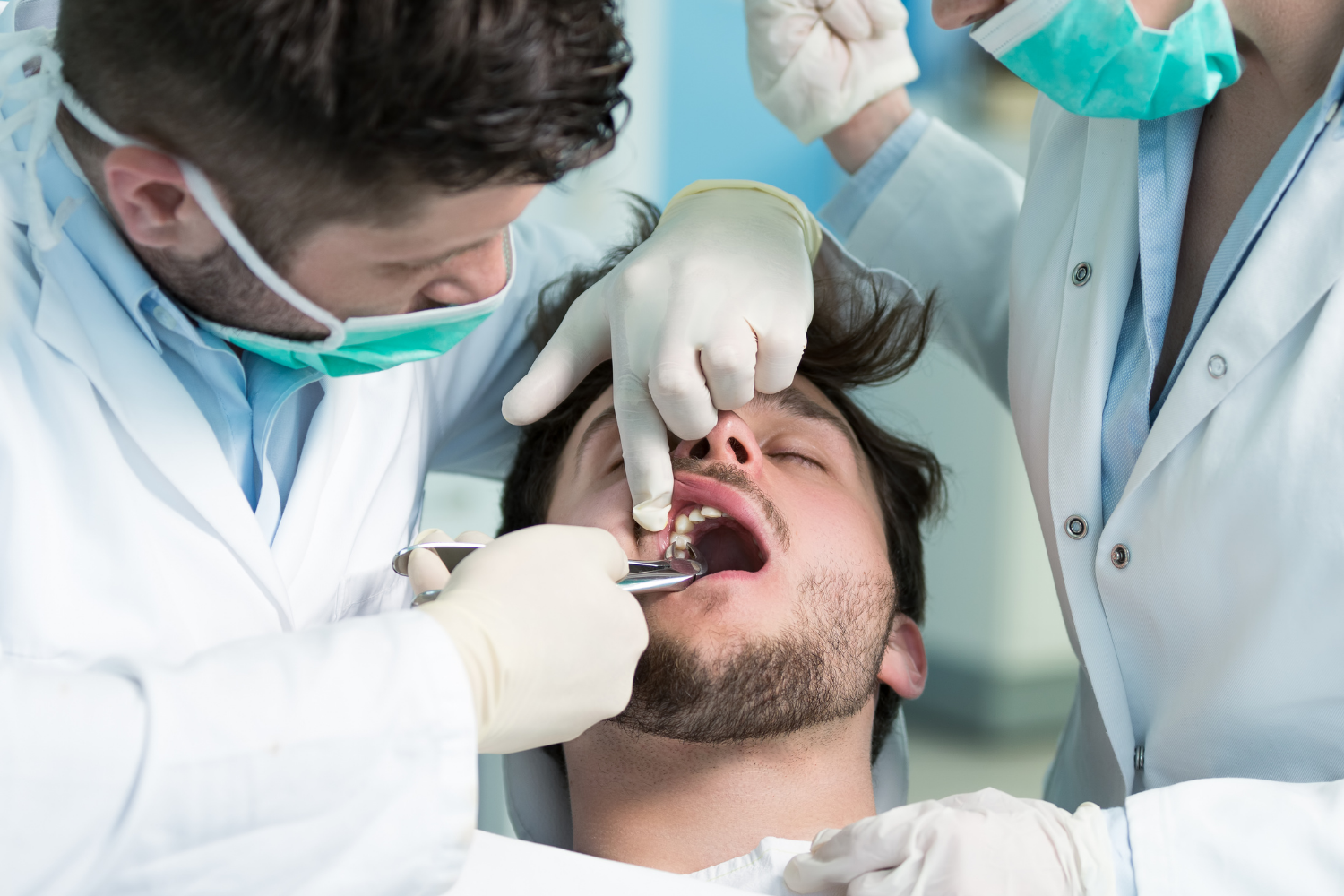 Dentist examining a patient’s mouth with two assistants in a clinic, all wearing masks and gloves