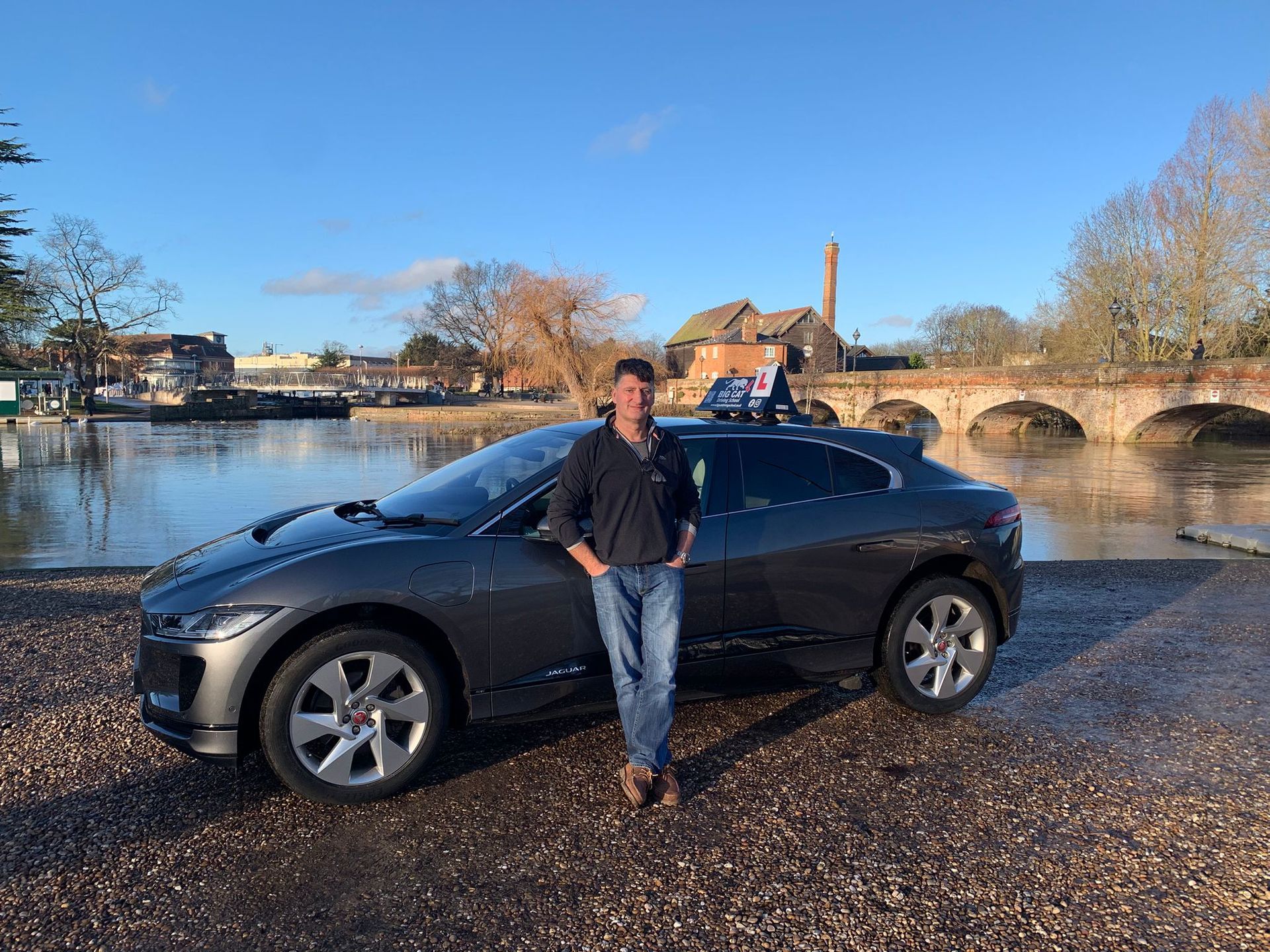 Man standing next to a grey Jaguar I-PACE by a river, with a bridge and buildings in the background.