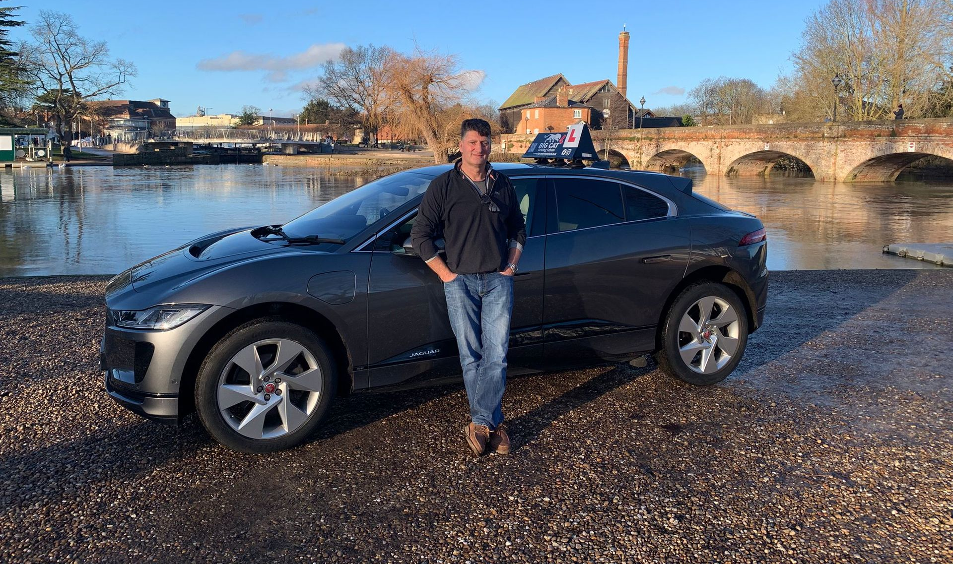 Man standing by a gray Jaguar I-Pace near a body of water with a bridge in the background.