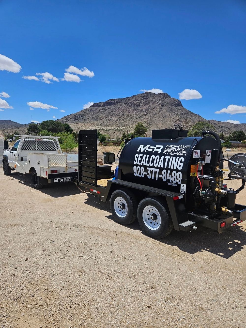 Two black utility trailers in a gravel lot with a mountain and blue sky in the background