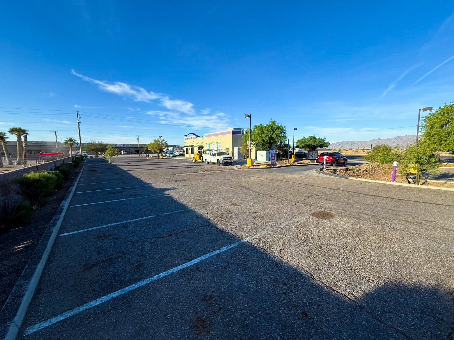 Empty gravel lot beside a road, with a small building and trees under a bright blue sky.