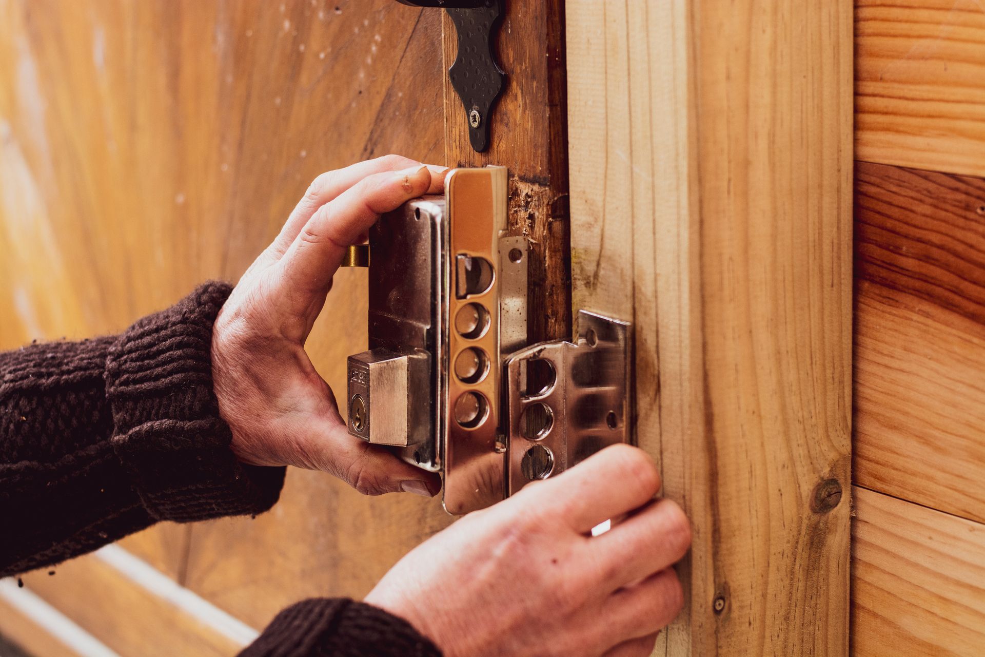 A person installs a shiny metal door lock on a wooden door with visible screws and a latch mechanism.