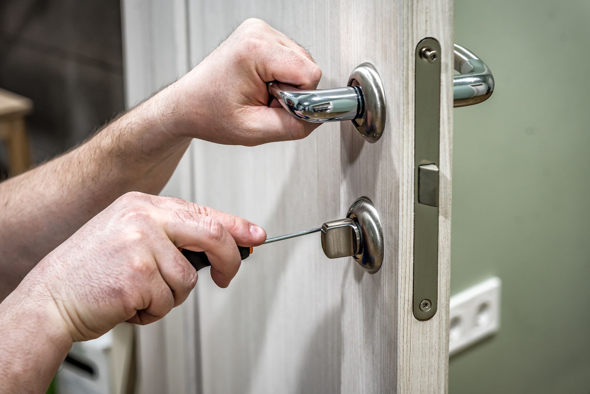 Hands replacing door lock using screwdriver on light wood door with metal handle.